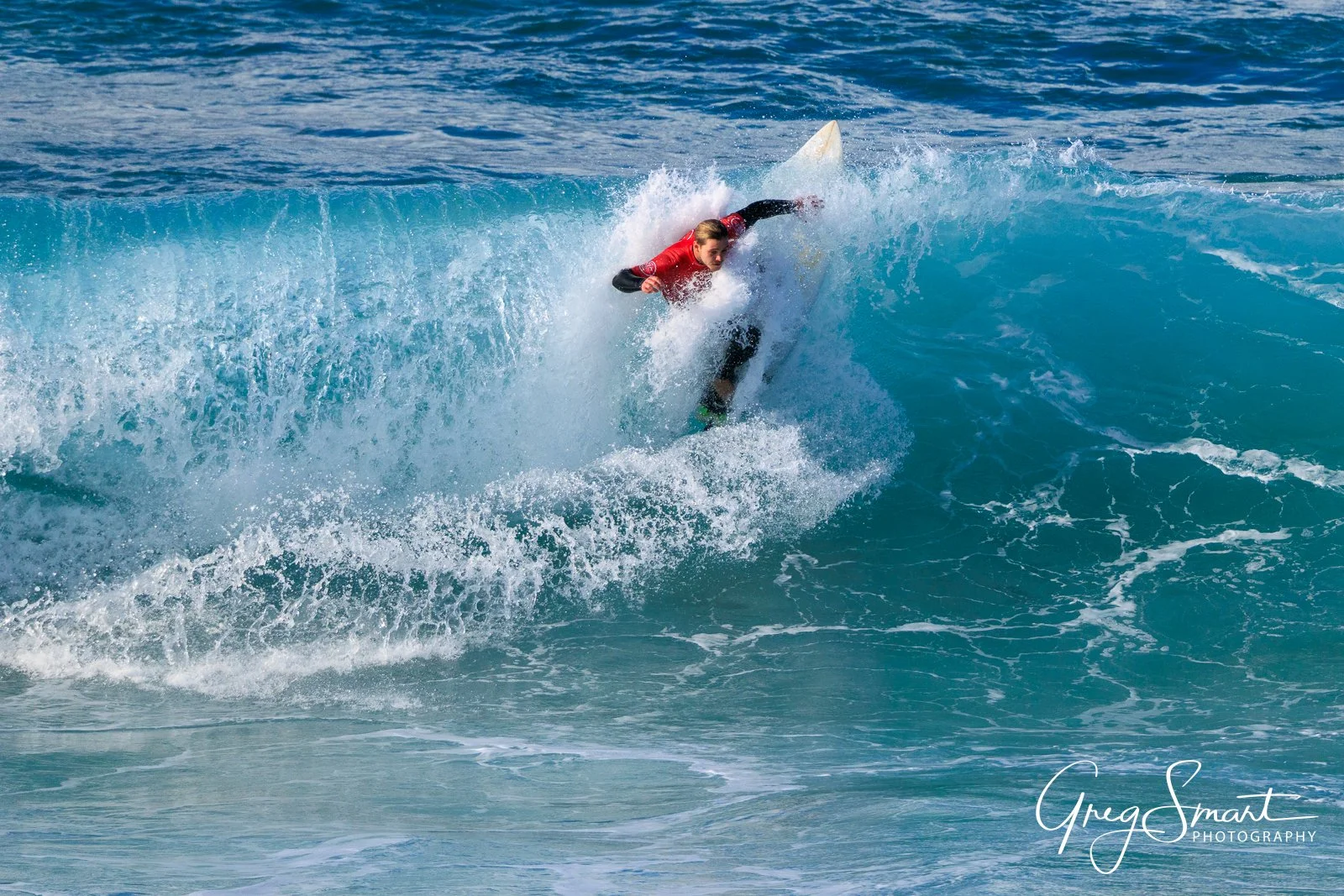 A person surfing on a blue wave in the ocean, wearing a red wetsuit top and black wetsuit bottom.