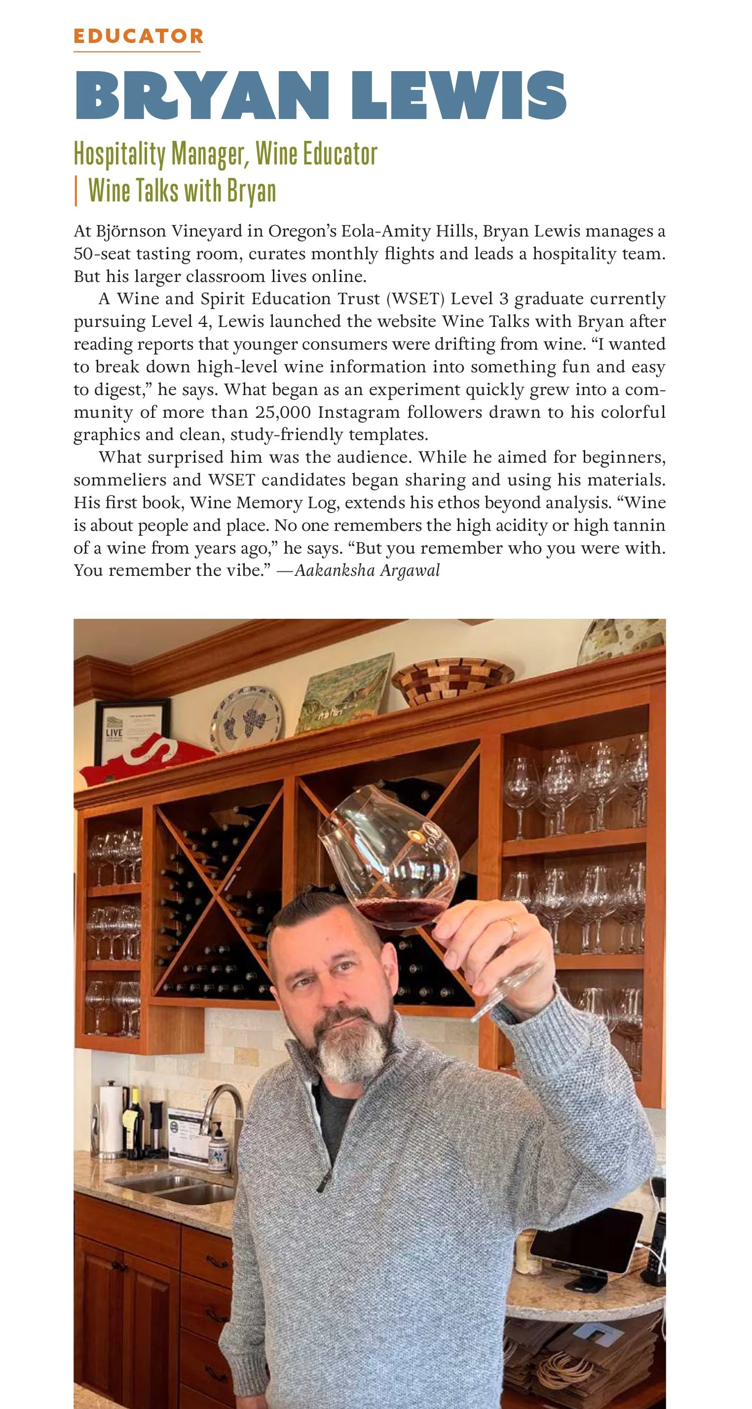 A man with a beard and mustache, wearing a gray sweater, holds a glass of red wine up in a kitchen with wooden cabinets, wine bottles, glasses, and decorative plates on the wall.