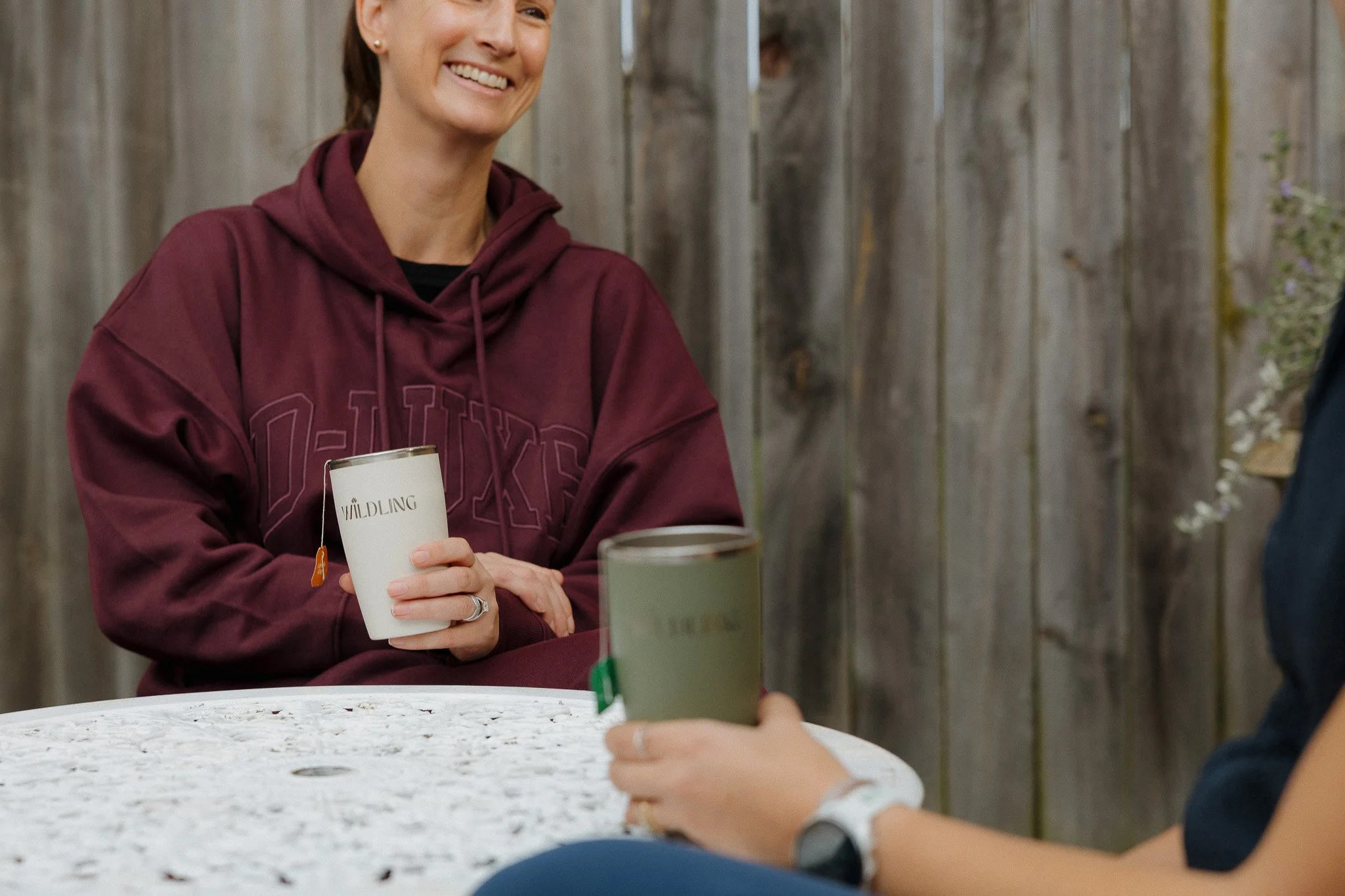 A woman in a maroon hoodie smiling and holding a white cup, sitting at a round table with a white cloth, with another person partially visible holding a similar cup in front of her. The background consists of a wooden fence.