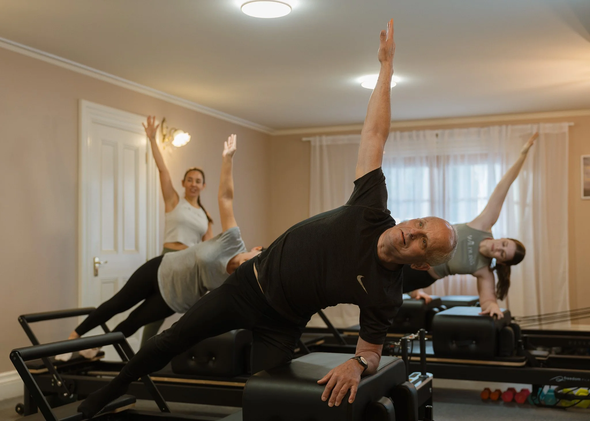 Group of people participating in a Pilates class in a home fitness room, with some lying on Pilates reformer machines performing exercises and a woman standing and smiling at the back.