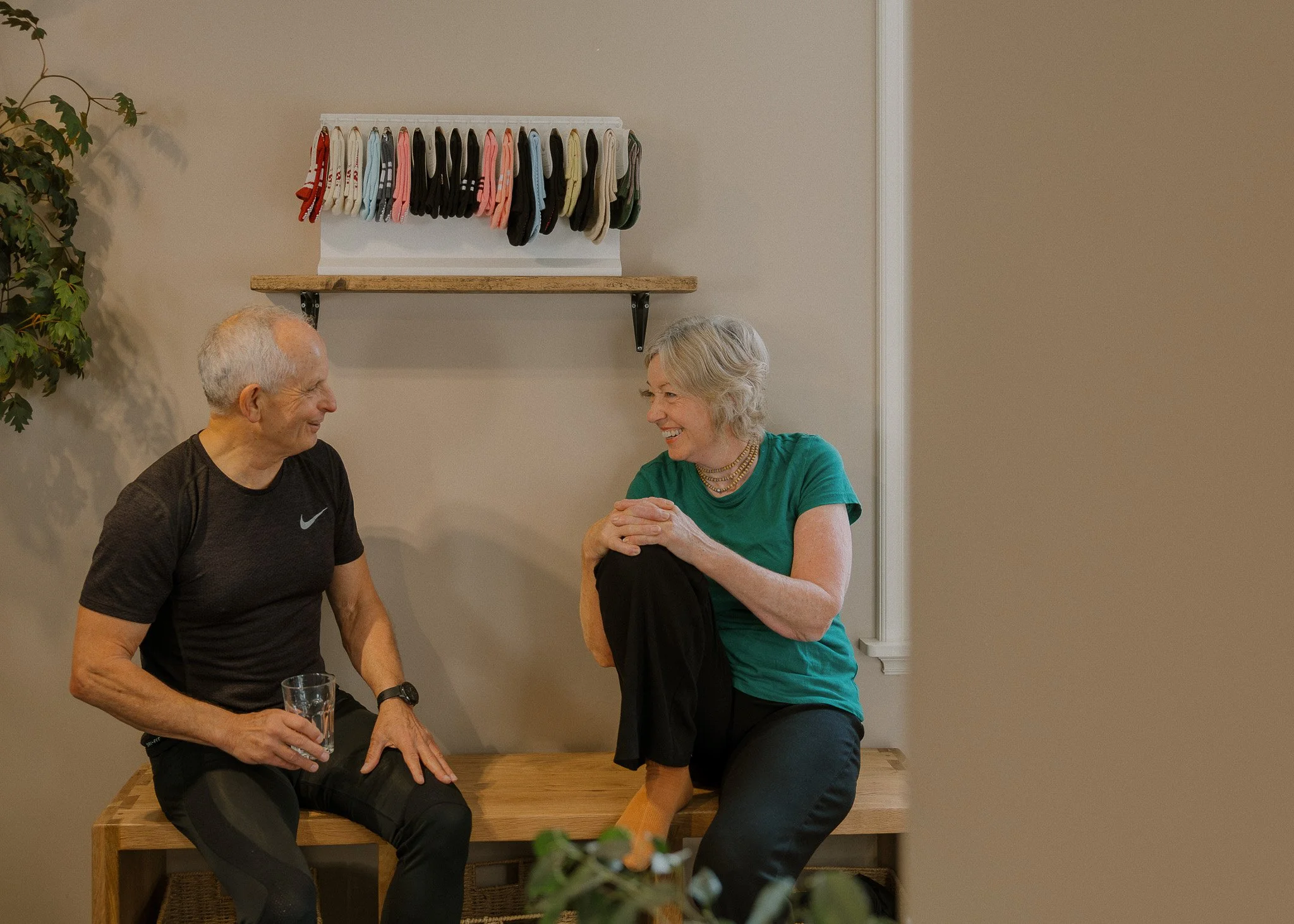 An elderly man and woman sitting on a wooden table, laughing and talking. The man is holding a glass of water, and they are engaging in a cheerful conversation. In the background, a rack of colorful socks hangs on the wall, next to a green potted plant.