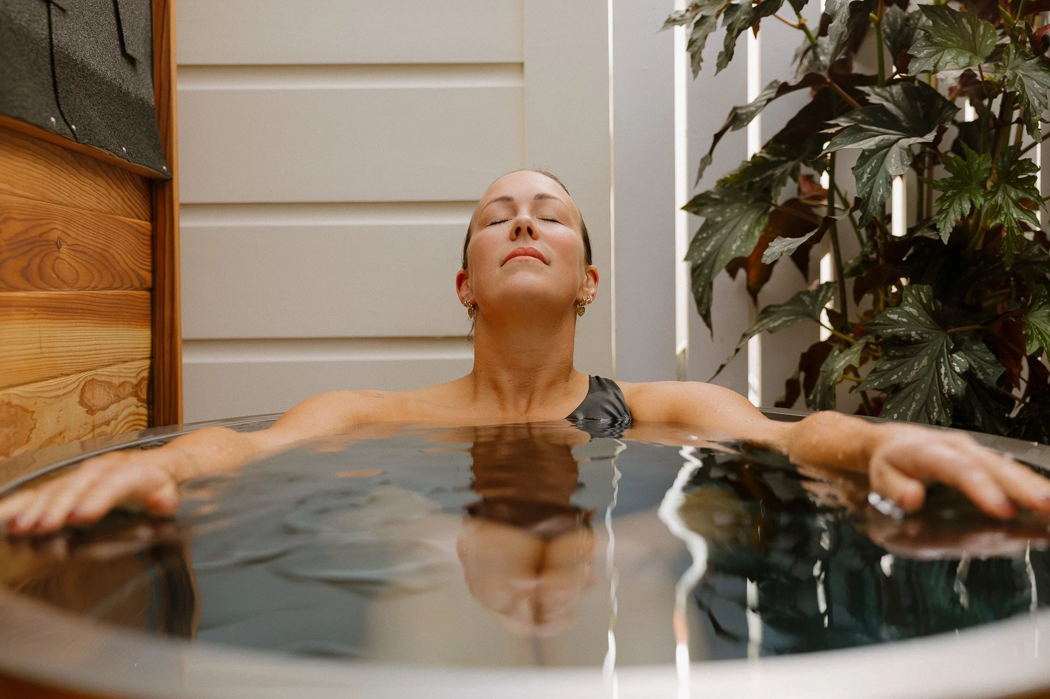 A woman with closed eyes and earrings relaxes in a hot tub, with her arms extended and water rippling around her. She is positioned outdoors next to a wooden wall and green foliage.
