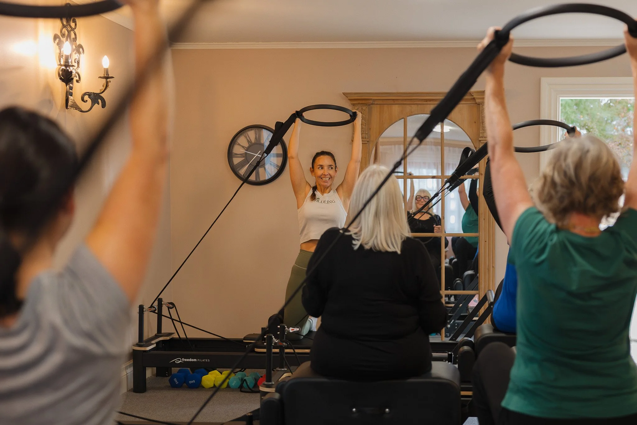 A group of people participating in a Pilates class using resistance rings in a studio, with a smiling instructor leading the session.