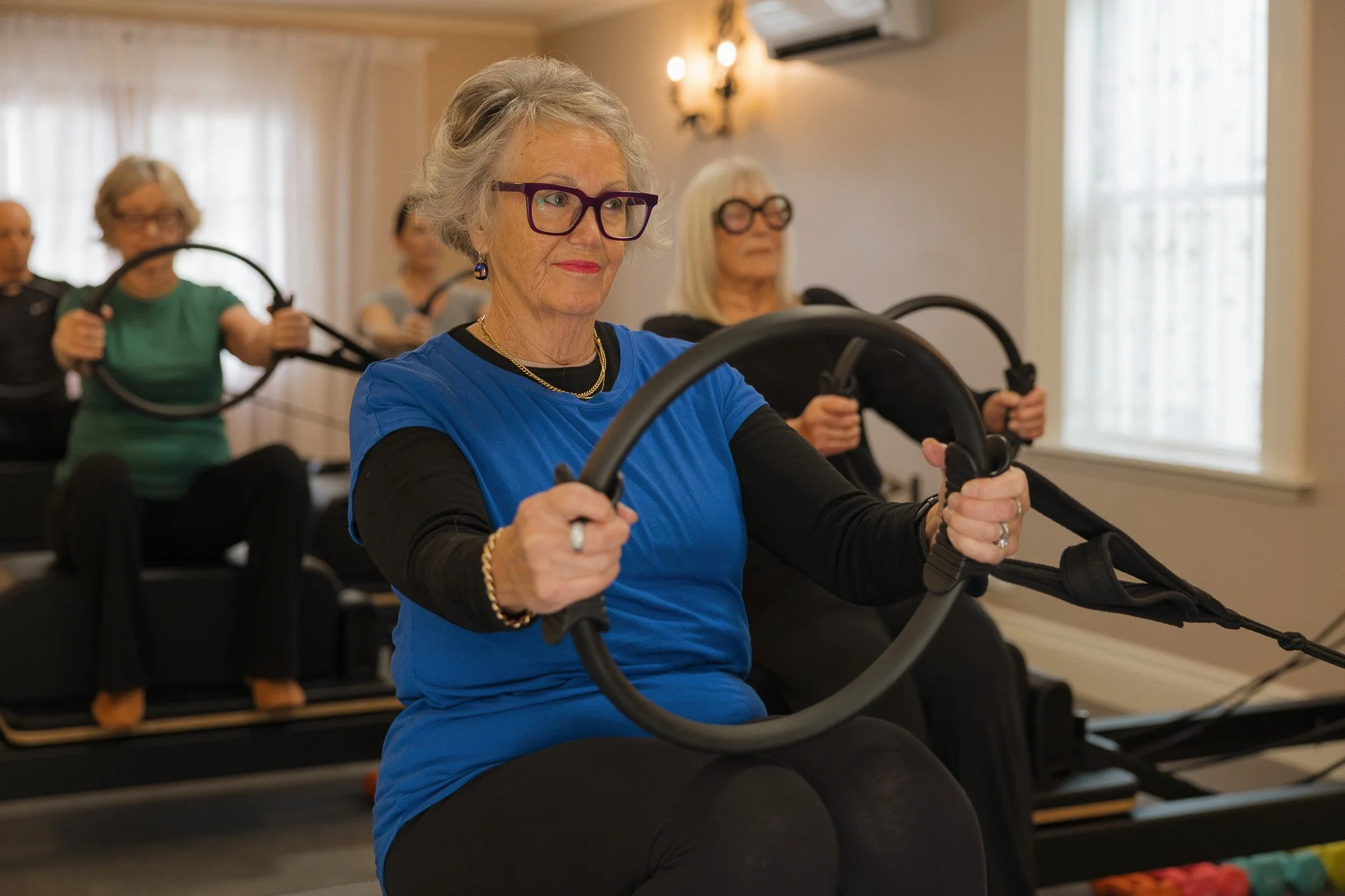 Older woman with gray hair, glasses, wearing a blue top, participating in a seated exercise class in a well-lit room, holding a resistance ring with both hands, surrounded by other women doing similar exercises.