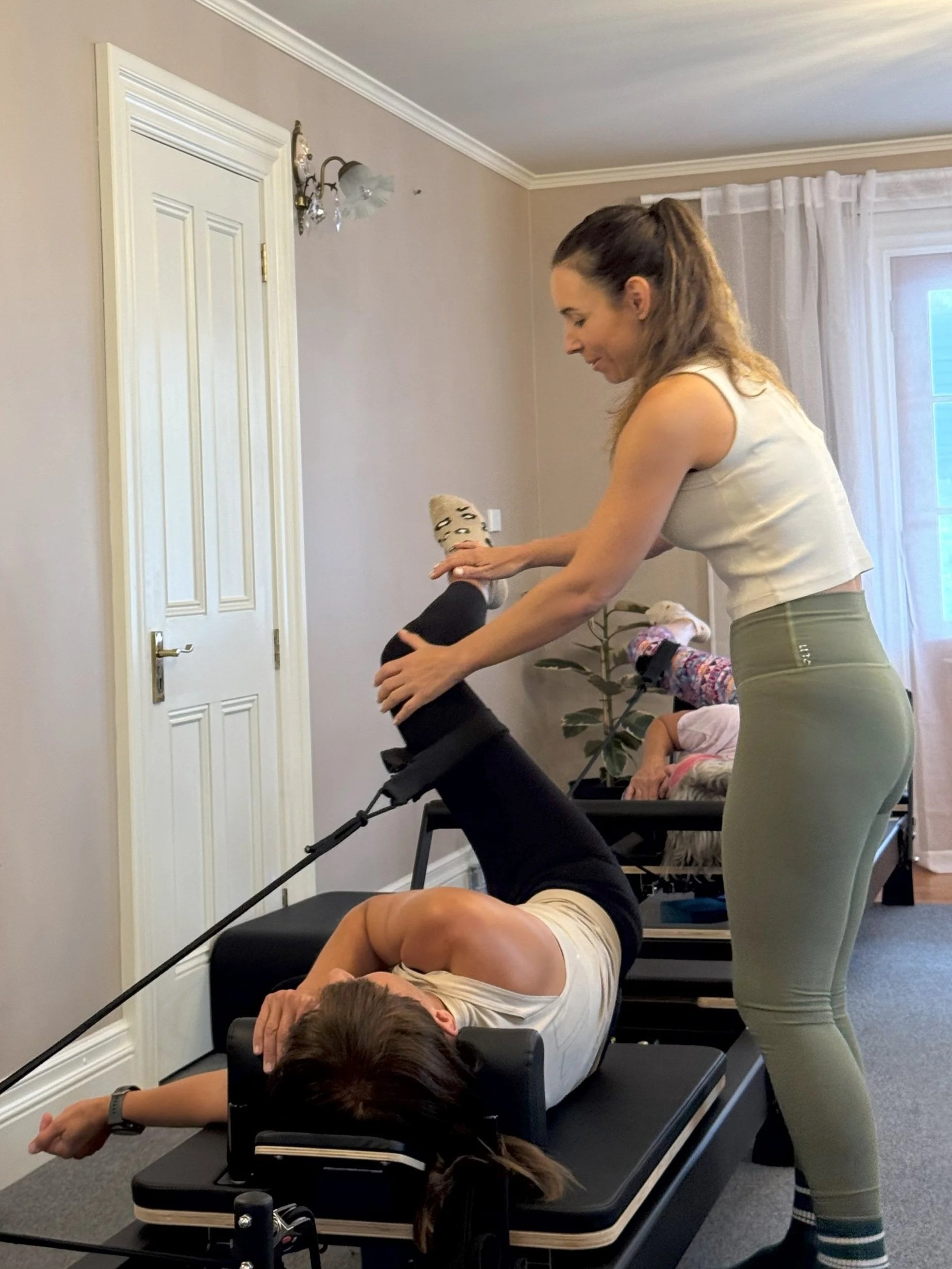 A woman assisting a person lying down. The woman is standing, wearing green leggings and a white tank top, and gently holding the person's legs. The person has their head resting on a padded surface, arms extended, and appears to be engaged in a physical exercise. There are other individuals in the background, indoor setting with a window and curtains.