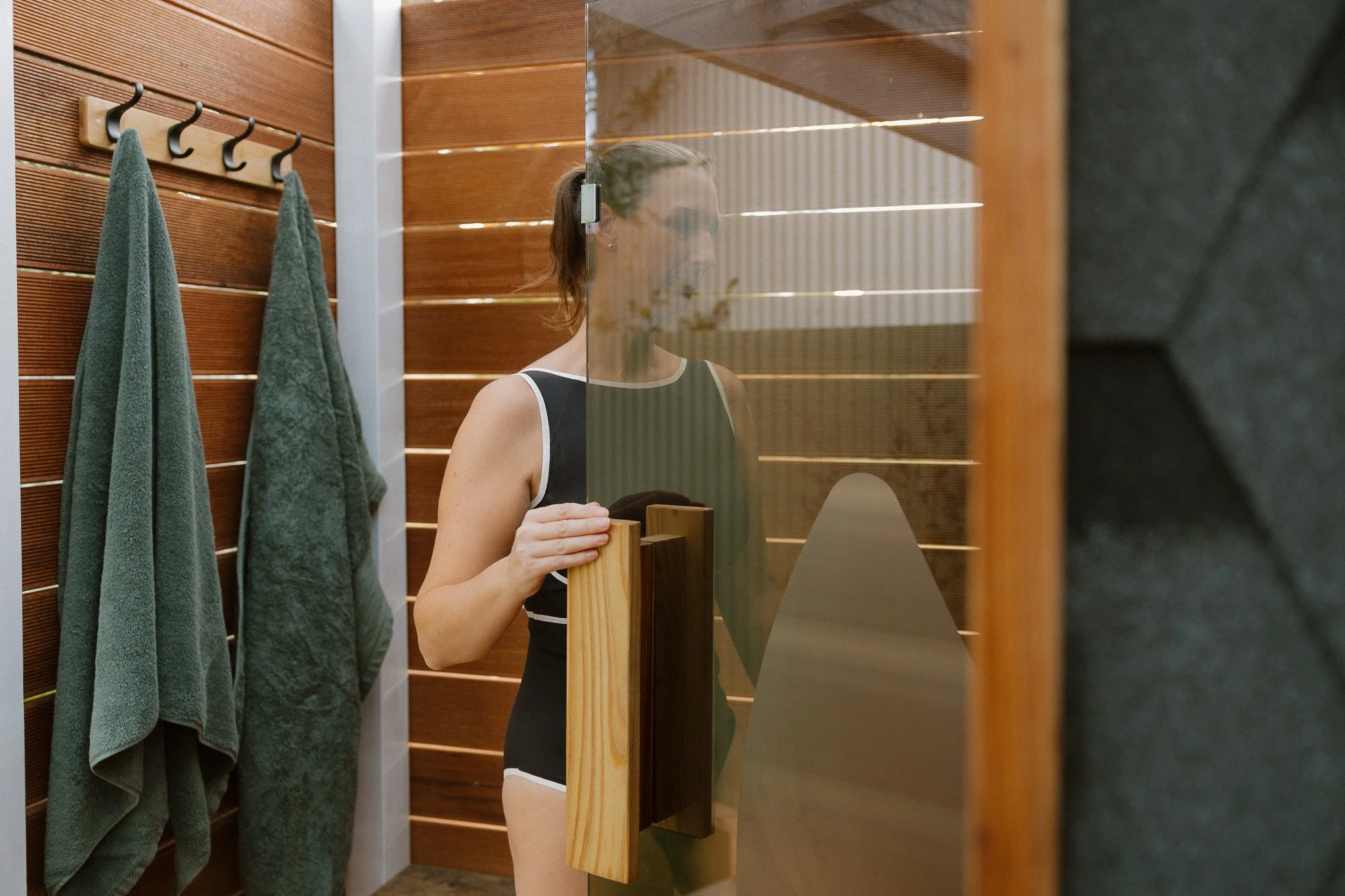 A woman looking in a mirror in a sauna or changing room, with green towels hanging on hooks on a wooden wall to her left.