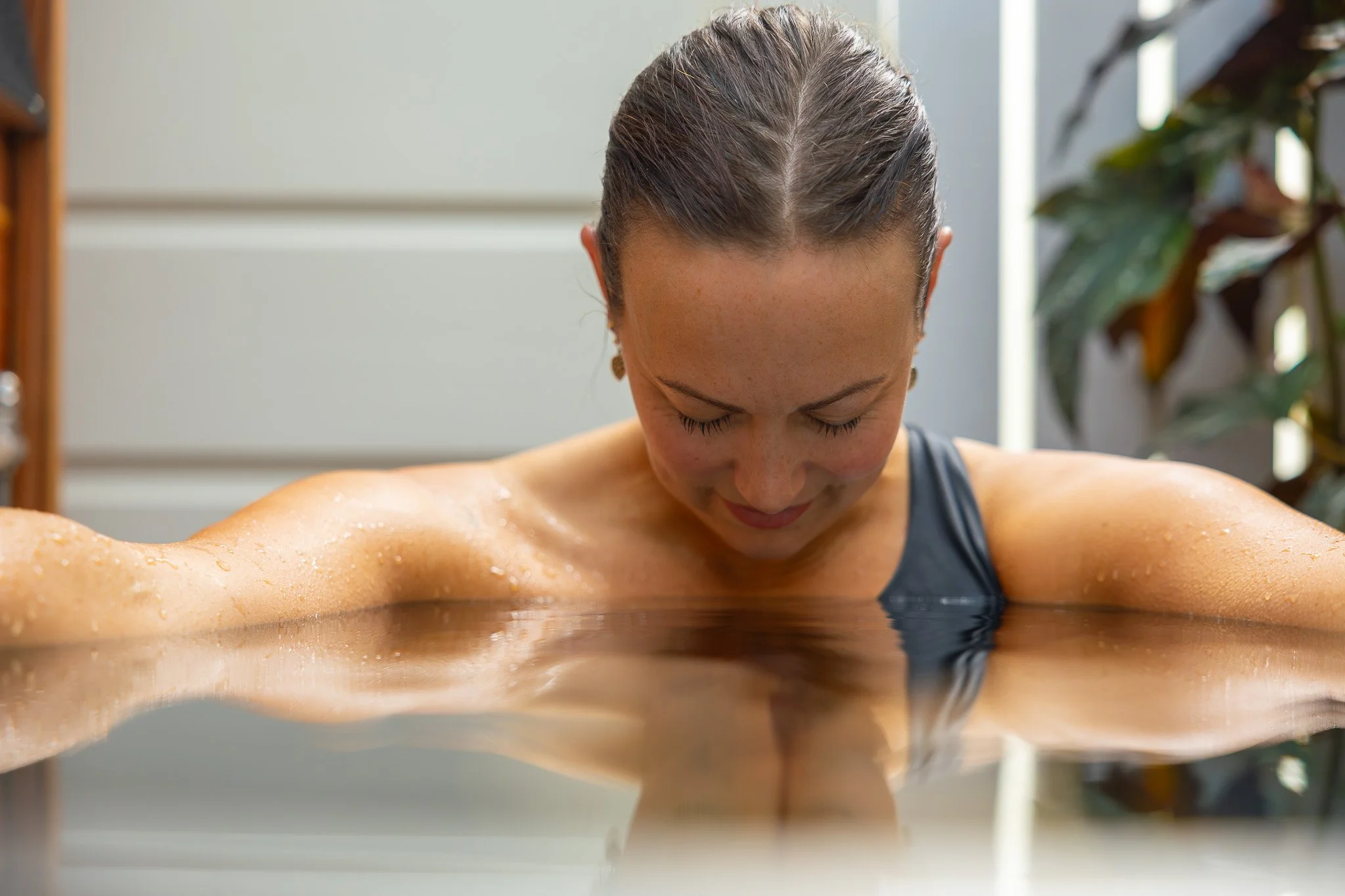 A woman with brown hair pulled back in a bun is leaning forward with her head down near a wooden surface, possibly a table or counter, with water or sweat on her shoulders and arms. She appears to be in a spa or wellness setting surrounded by indoor plants.