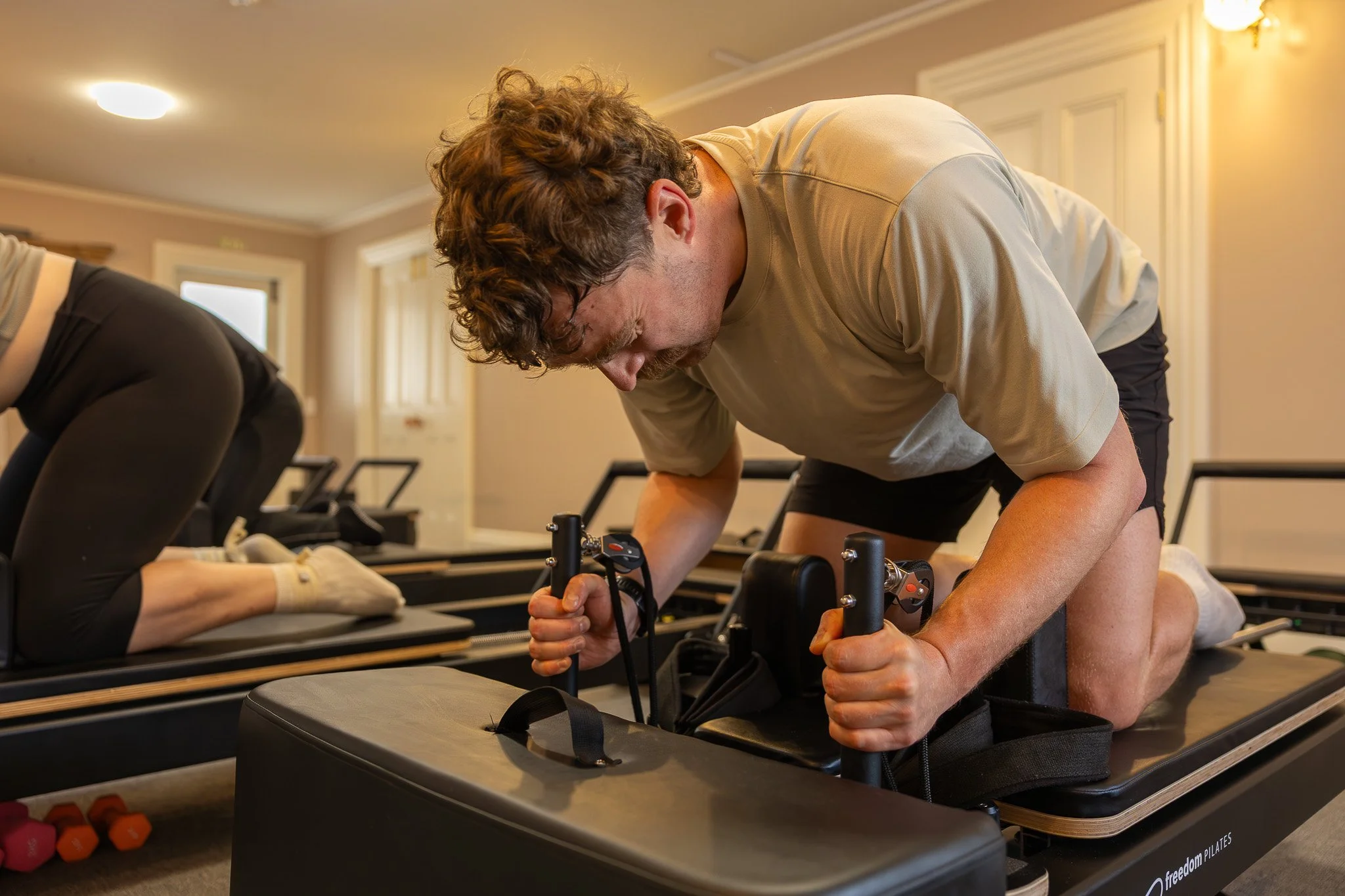 A man with curly brown hair and a mustache performing a Pilates exercise on a reformer machine in a fitness studio, with another person next to him partially visible.