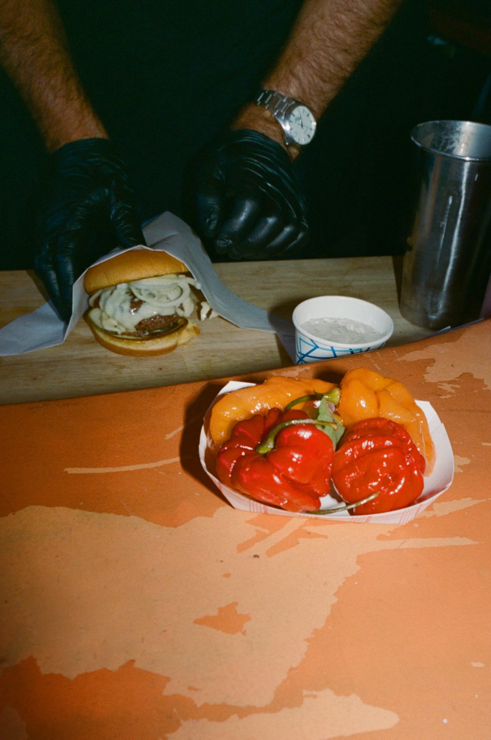 Person wearing black gloves and a watch preparing a cheeseburger with onions. In the foreground, there is a bowl of red and orange bell peppers on an orange surface.