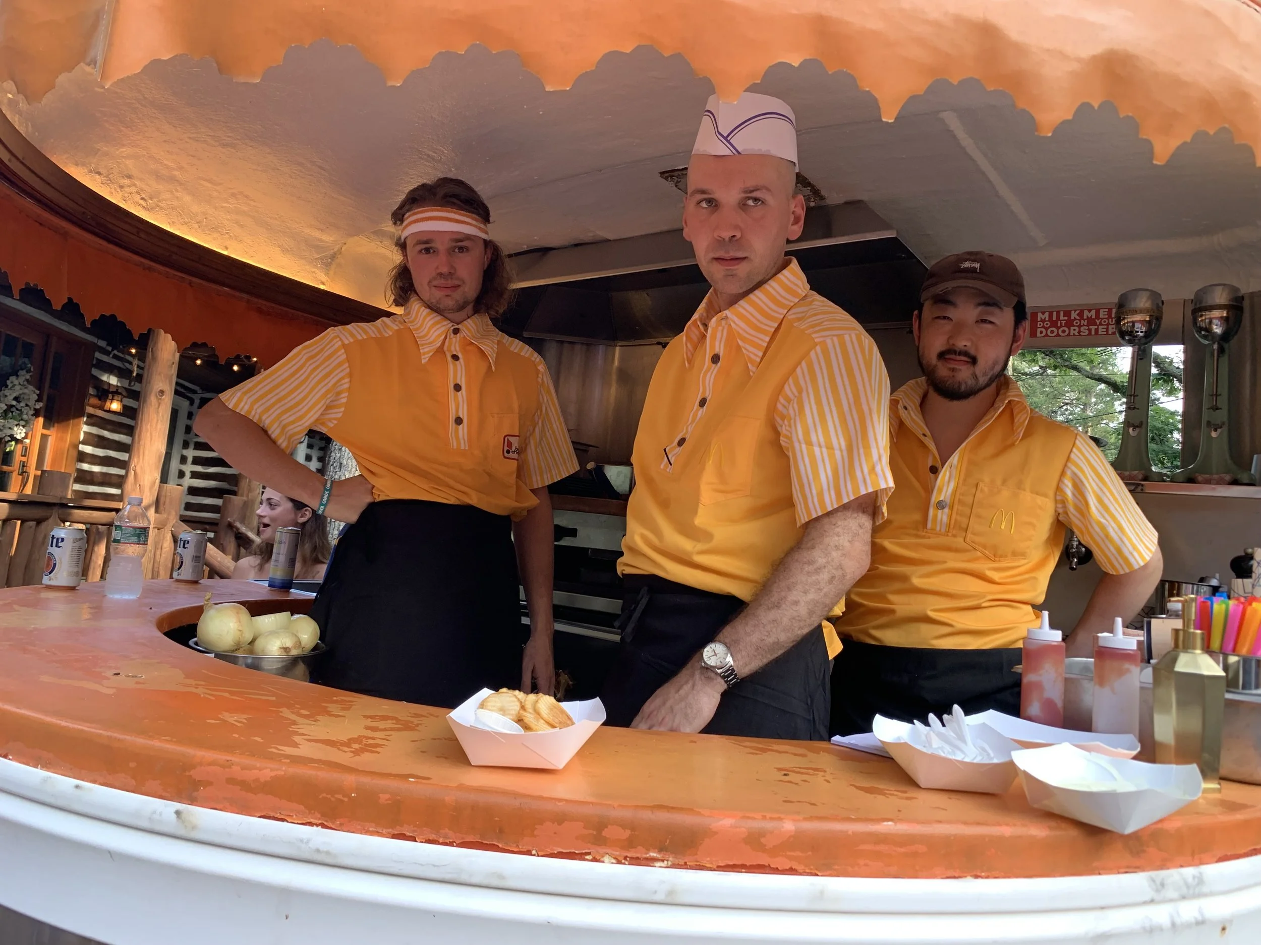 Three McDonald's employees in yellow uniforms working at a food stand, with one person wearing a striped headband and another wearing a hat, on a restaurant or food stall.