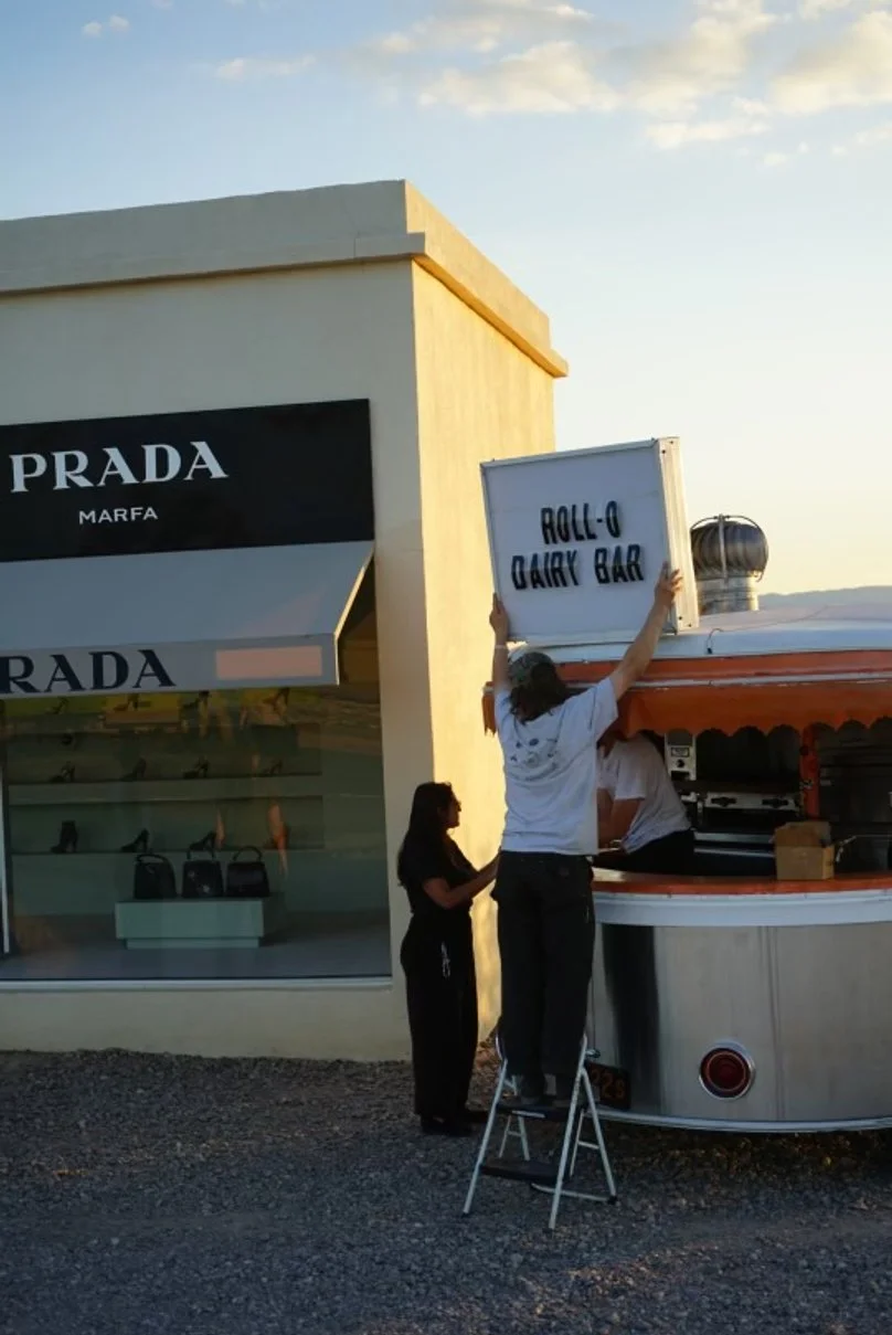 People setting up a sign that reads 'Roll-Q Dairy Bar' outside near a Prada store during sunset.