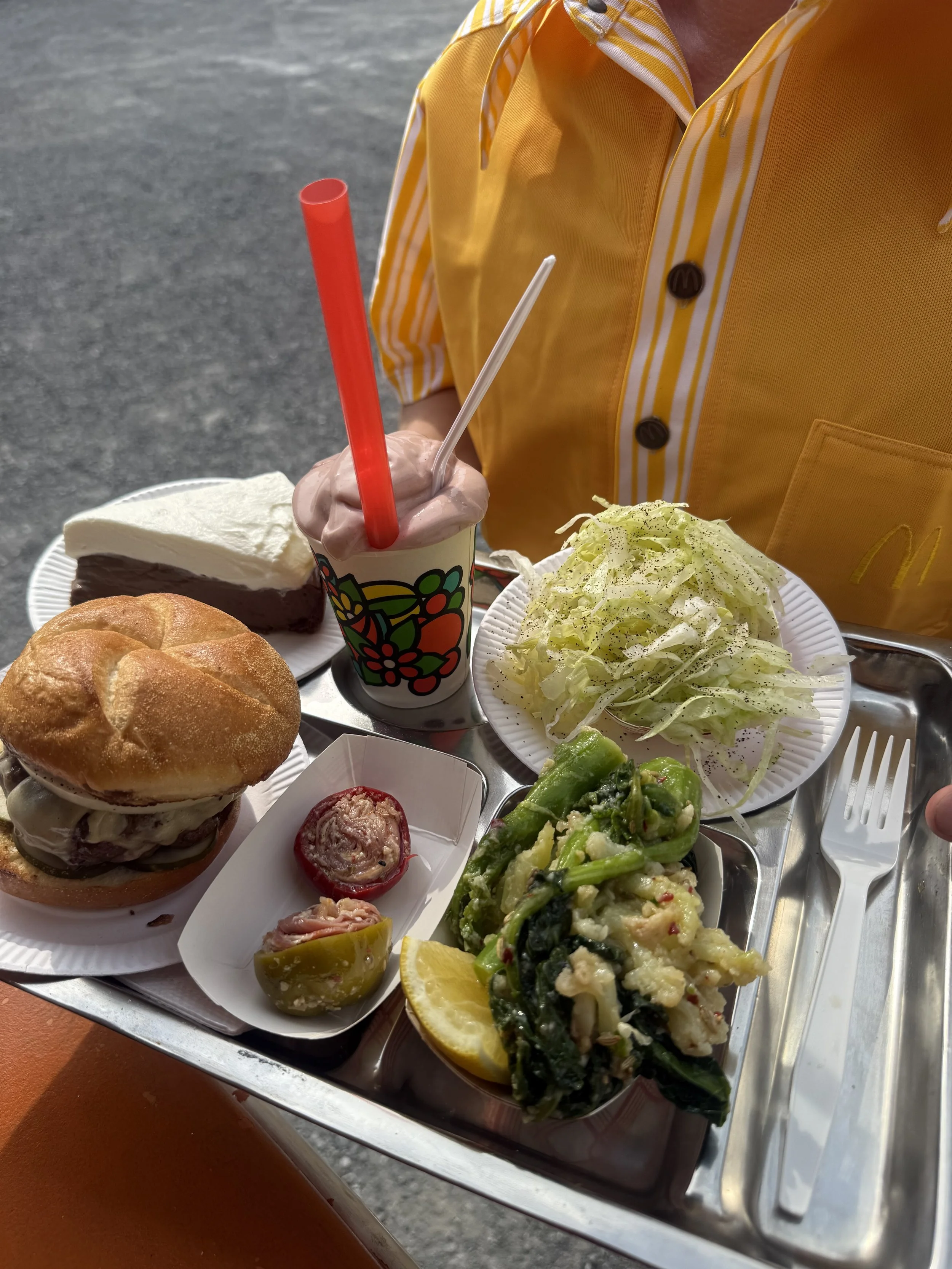 Food tray with burger, salad, pickles, lemon wedge, cake, ice cream, and a milkshake, held by a person wearing a yellow striped shirt.