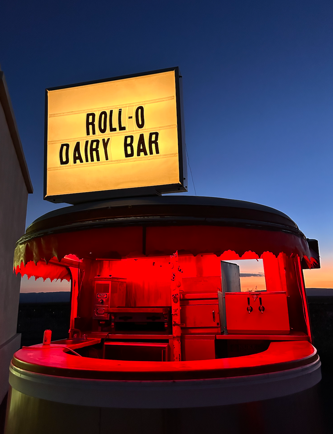 A food cart with a sign that reads "Rollo Dairy Bar" against a backdrop of a blue sky at sunset.