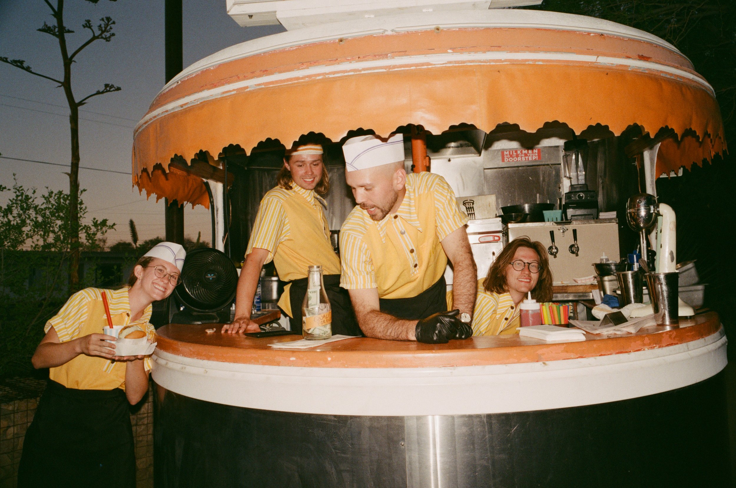 Four people dressed in yellow and white striped uniforms working inside a retro-style ice cream stand at sunset.