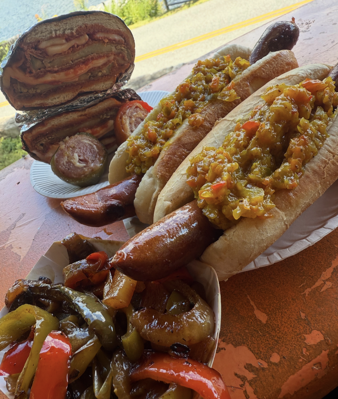 Hot dogs topped with relish and mustard, a sausage link, grilled vegetables, and a hamburger with onions and pickles, all on a table outdoors.
