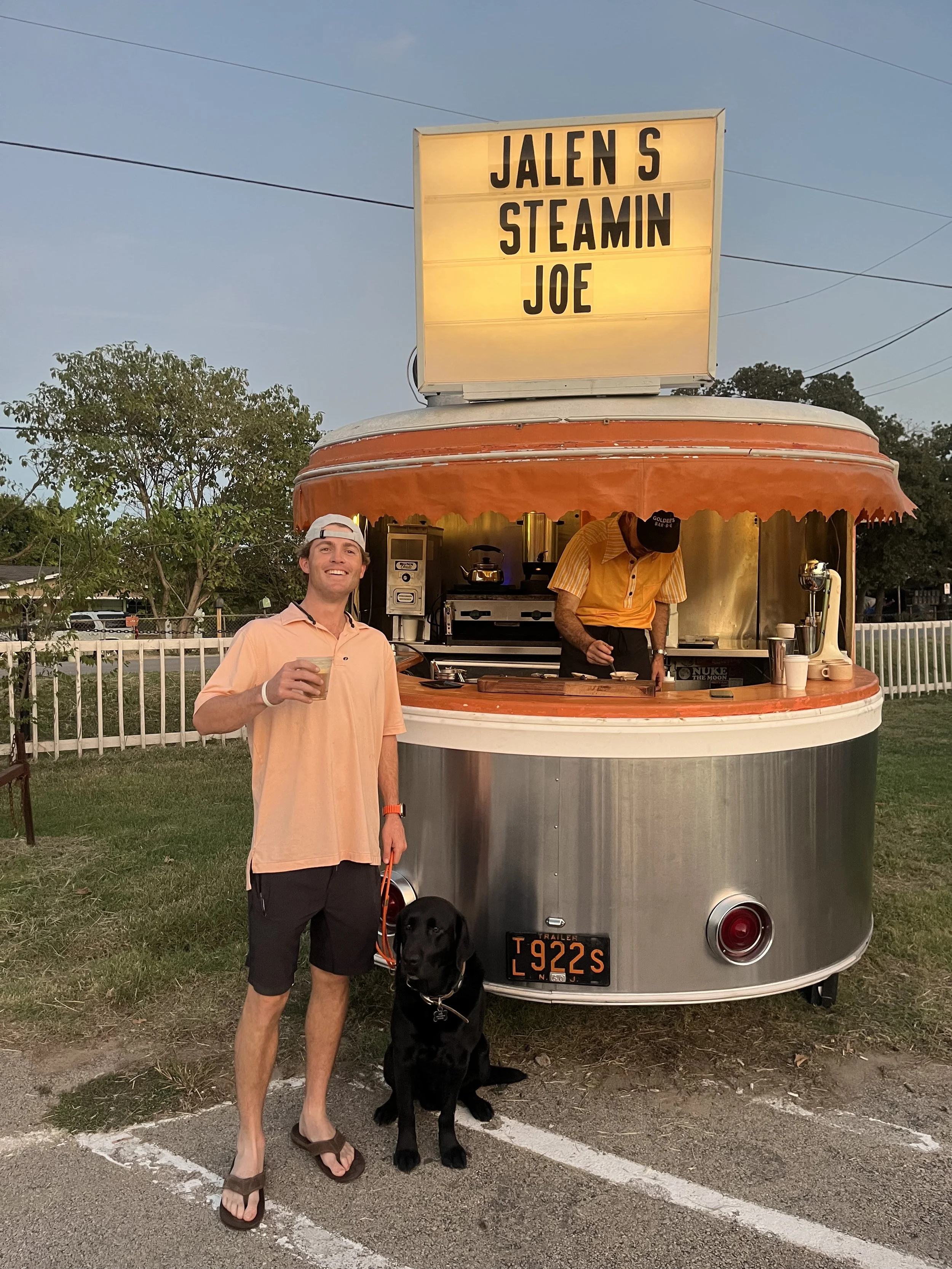 A happy man holding a drink standing next to a black labrador dog in front of a vintage food trailer with a sign that reads "JALEN S STEAMIN JOE." The trailer has an orange and silver exterior, and a person inside preparing food.