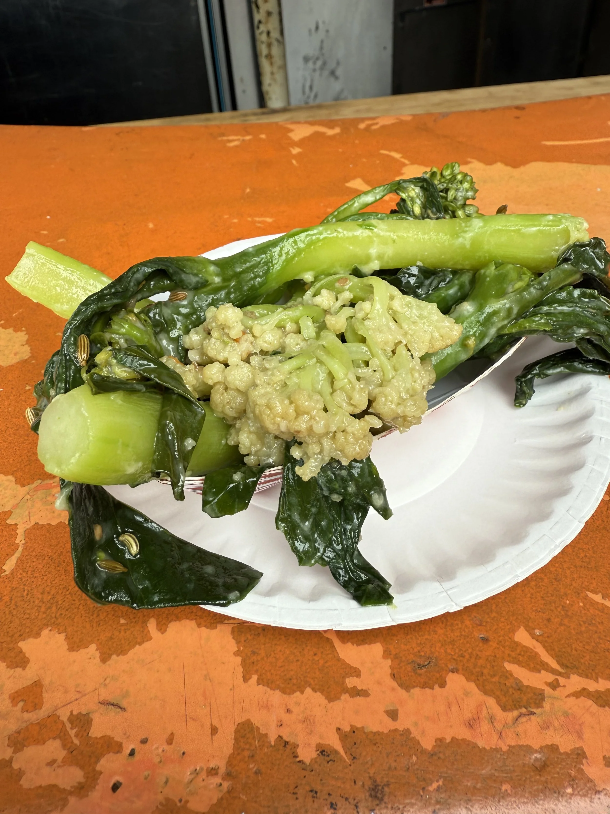 A serving of stir-fried leafy greens and vegetables on a white paper plate, placed on a worn orange surface.