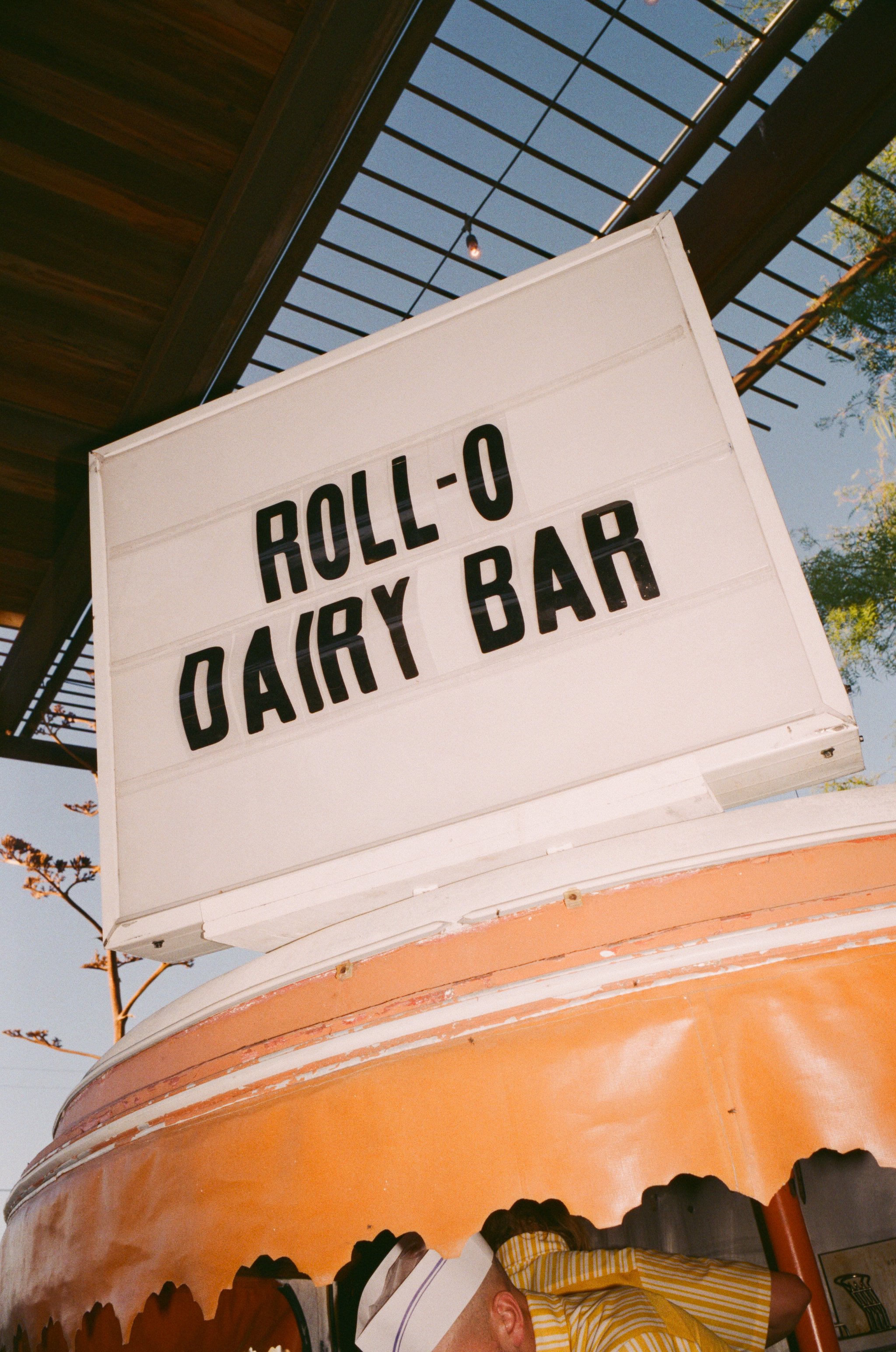 A sign displaying 'Roll-O Dairy Bar' outside a building with a wooden canopy and sky in the background.