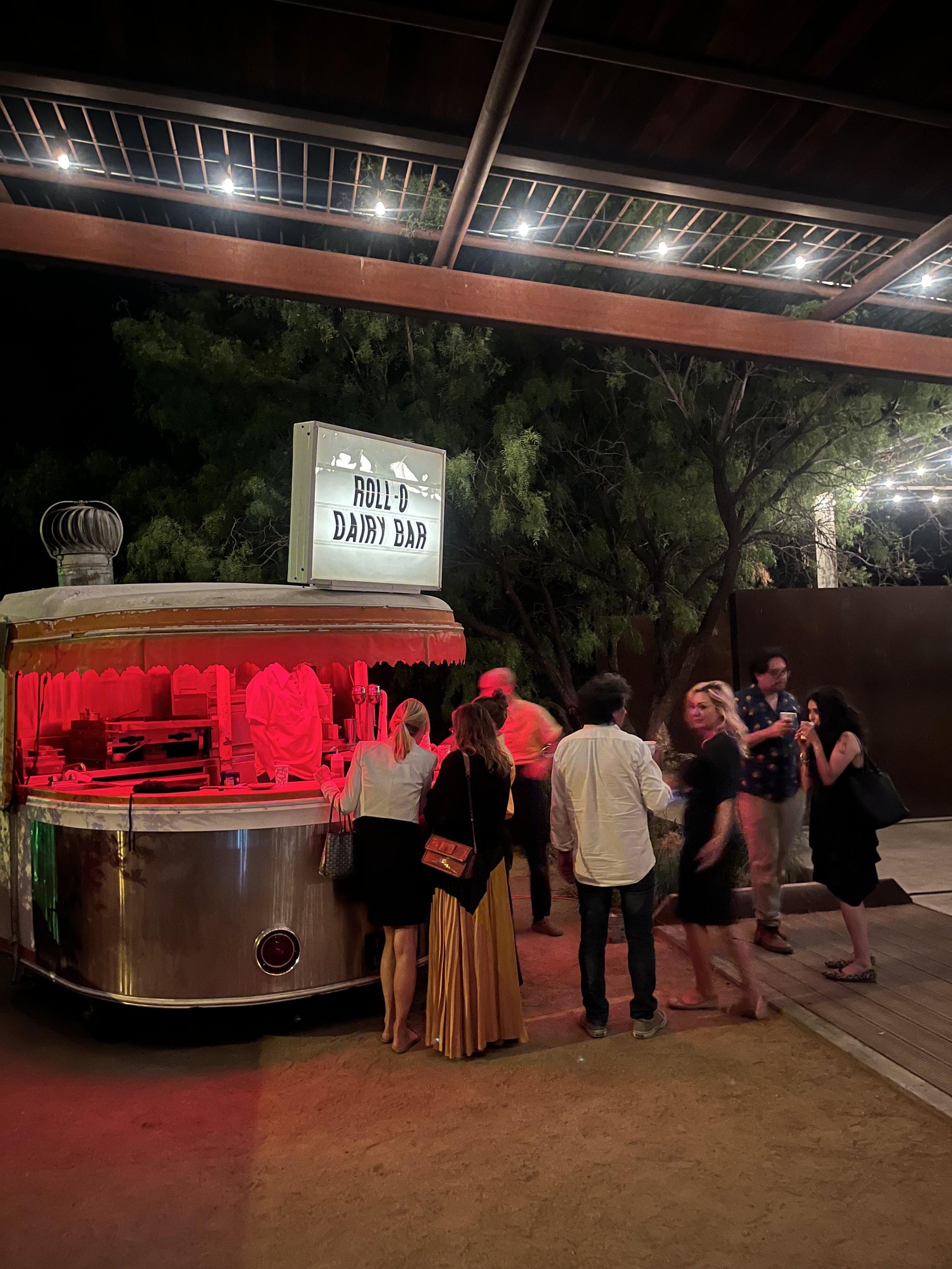 People gathered around a shiny, vintage-style mobile dairy bar food cart at night, with a sign reading 'Roll-O Dairy Bar' on top, under string lights and above a gravel surface.