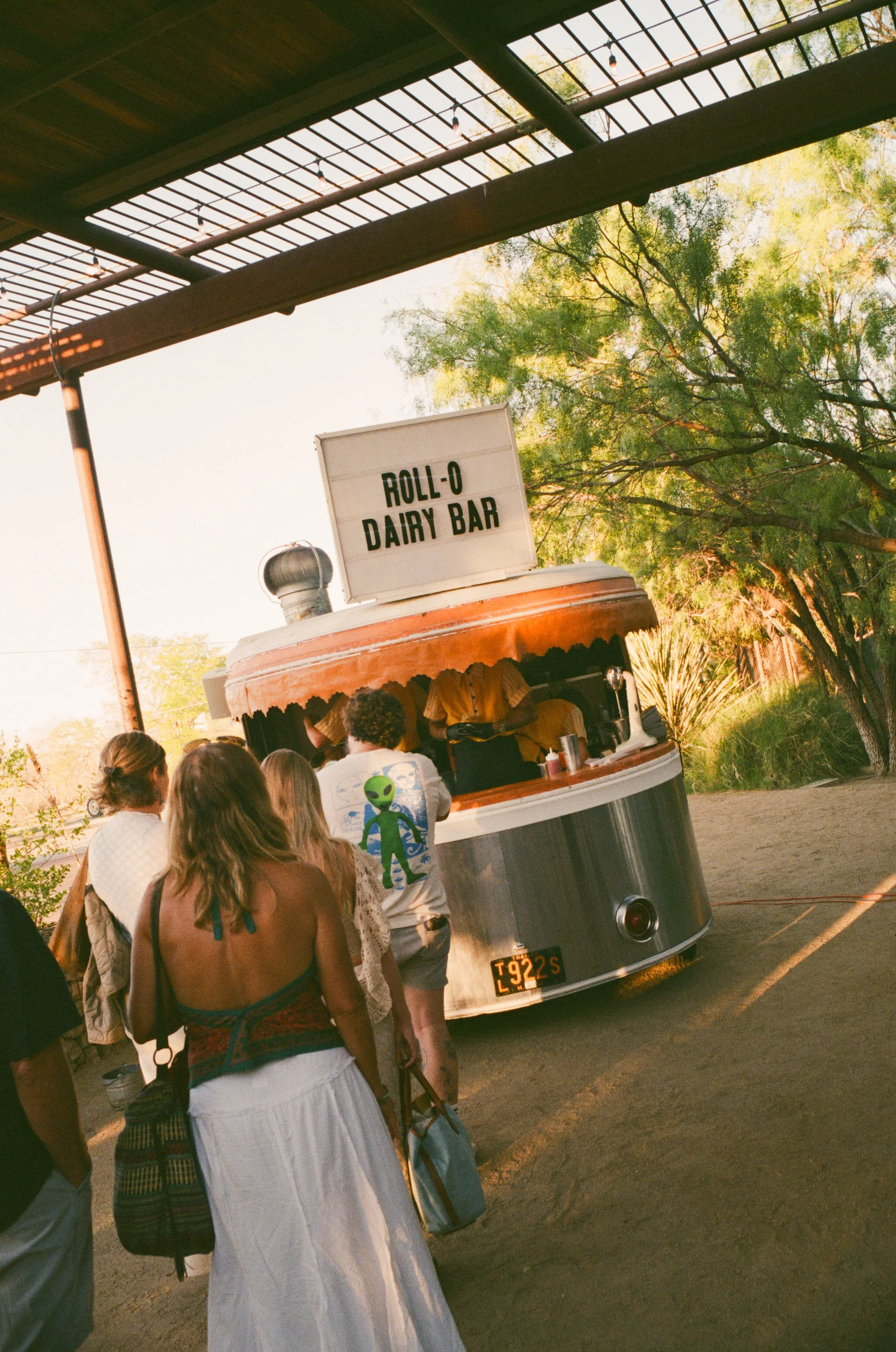 People standing in line order from a vintage-style, chrome dairy bar food truck with a sign that reads 'Roll-O Dairy Bar.' The truck is parked outdoors under a roof with trees in the background.