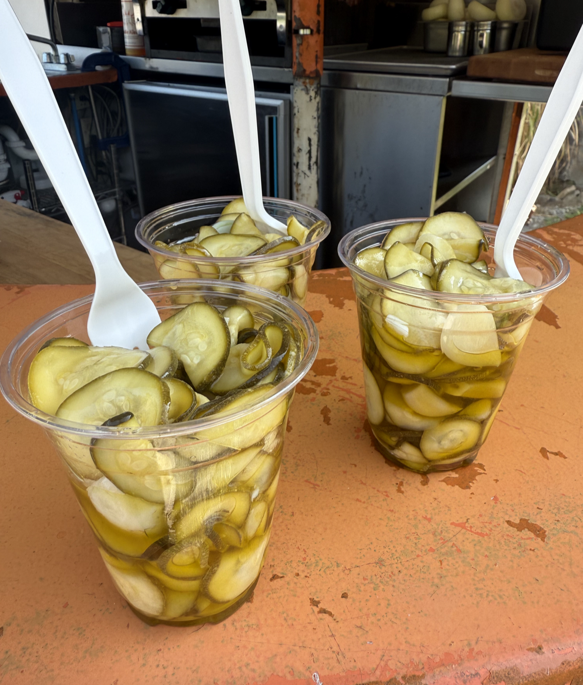 Three clear plastic cups filled with sliced pickles, each with a white plastic spoon inside, placed on an orange surface.