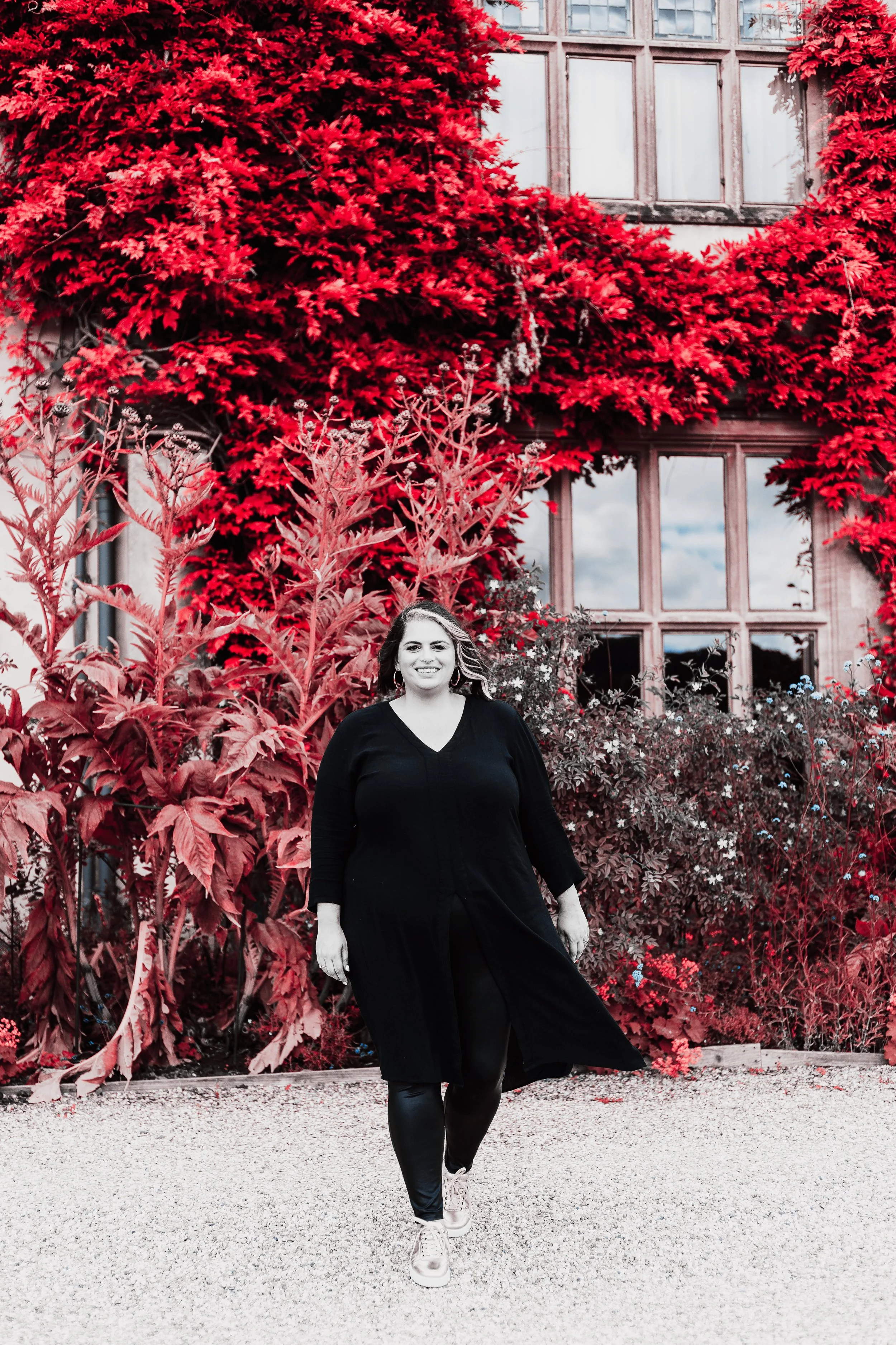 A woman with a smile, wearing a long black dress, white sneakers, and hoop earrings, walking outside in front of a background of red and green foliage and a building with large windows.