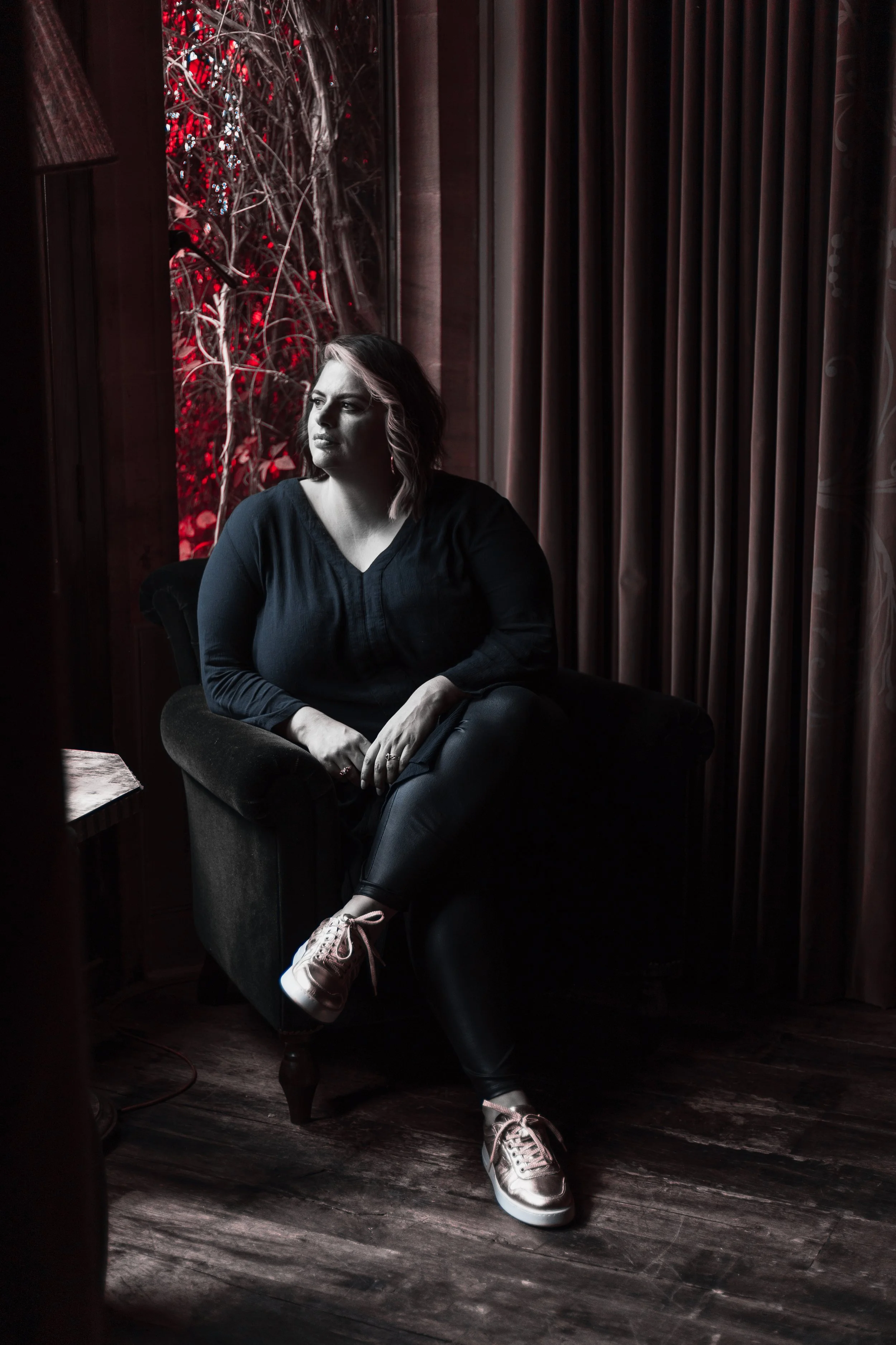 A woman with shoulder-length wavy hair, wearing a dark shirt and sneakers, sitting in a vintage armchair. She is indoors, next to a window with curtains, with a background of withered vines and red heat-sensitive lighting.
