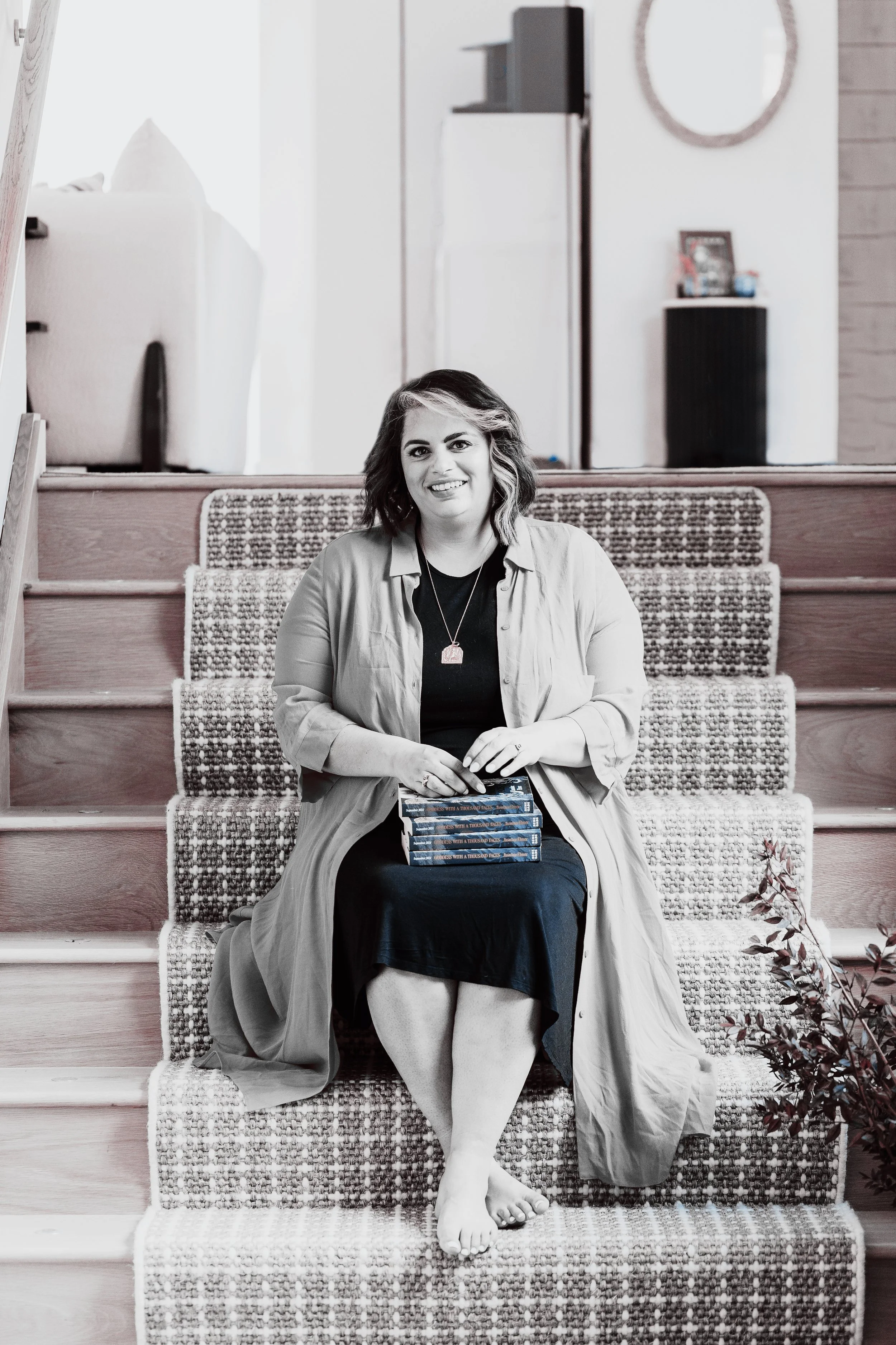 A woman sitting on carpeted stairs in a house, smiling, with a stack of books on her lap, wearing a long coat and dress, with scattered leaves or autumn decoration nearby.