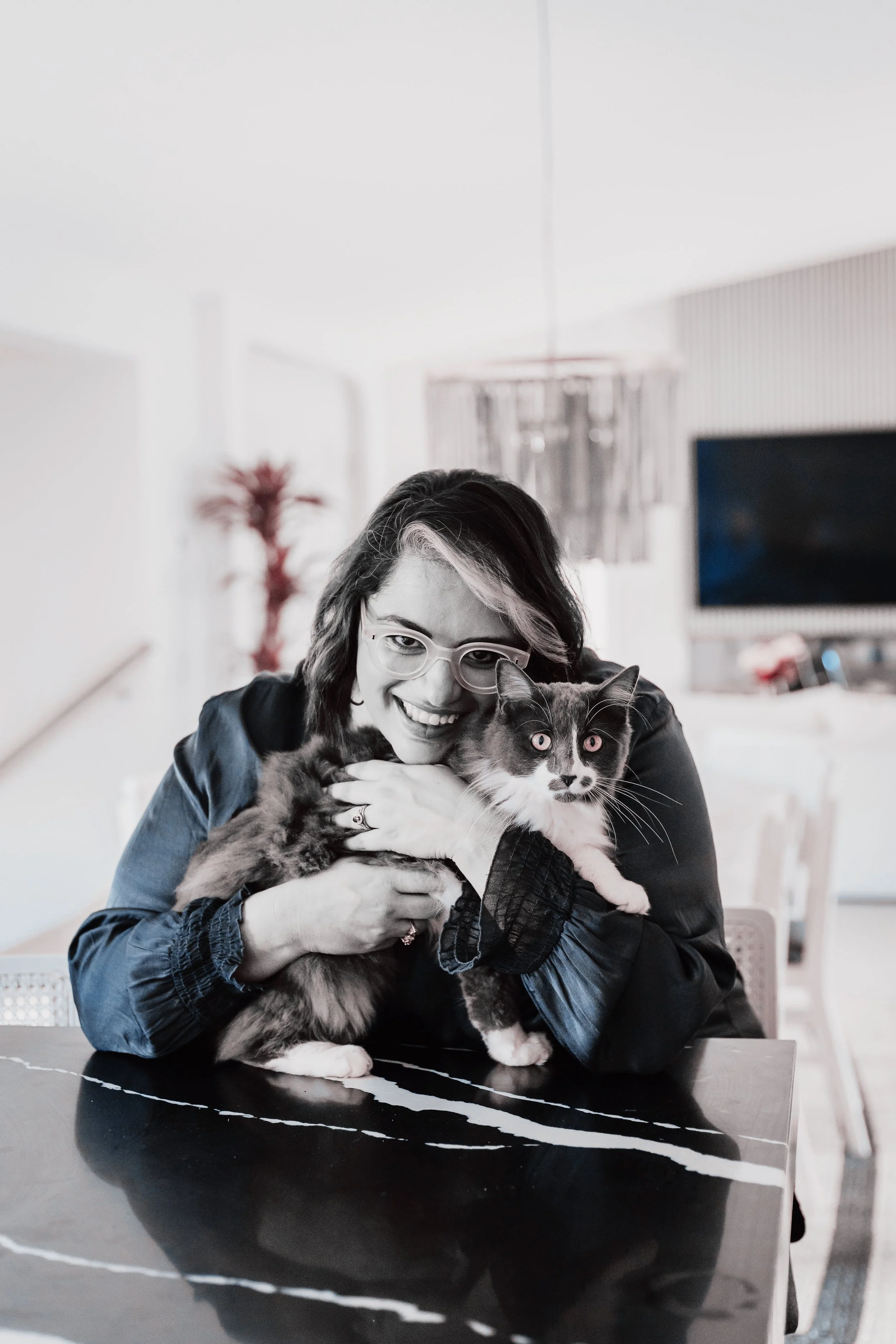 A woman with glasses and dark hair smiling while holding a gray and white cat in a modern, bright living room.