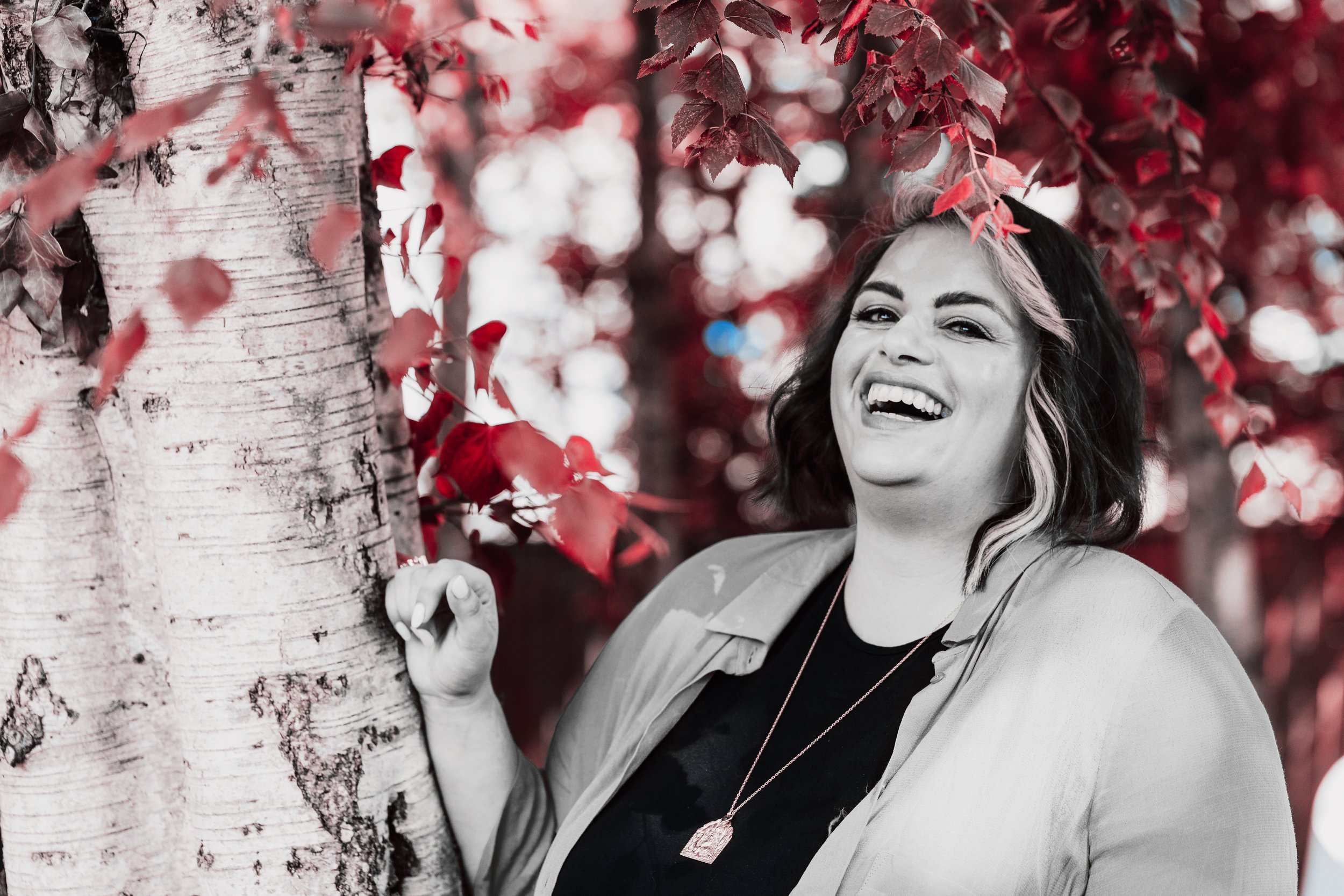 A woman with dark hair and a bright smile is standing beside a tree with reddish leaves, partly obscured by a black and white filter. She is outdoors in a park or garden.