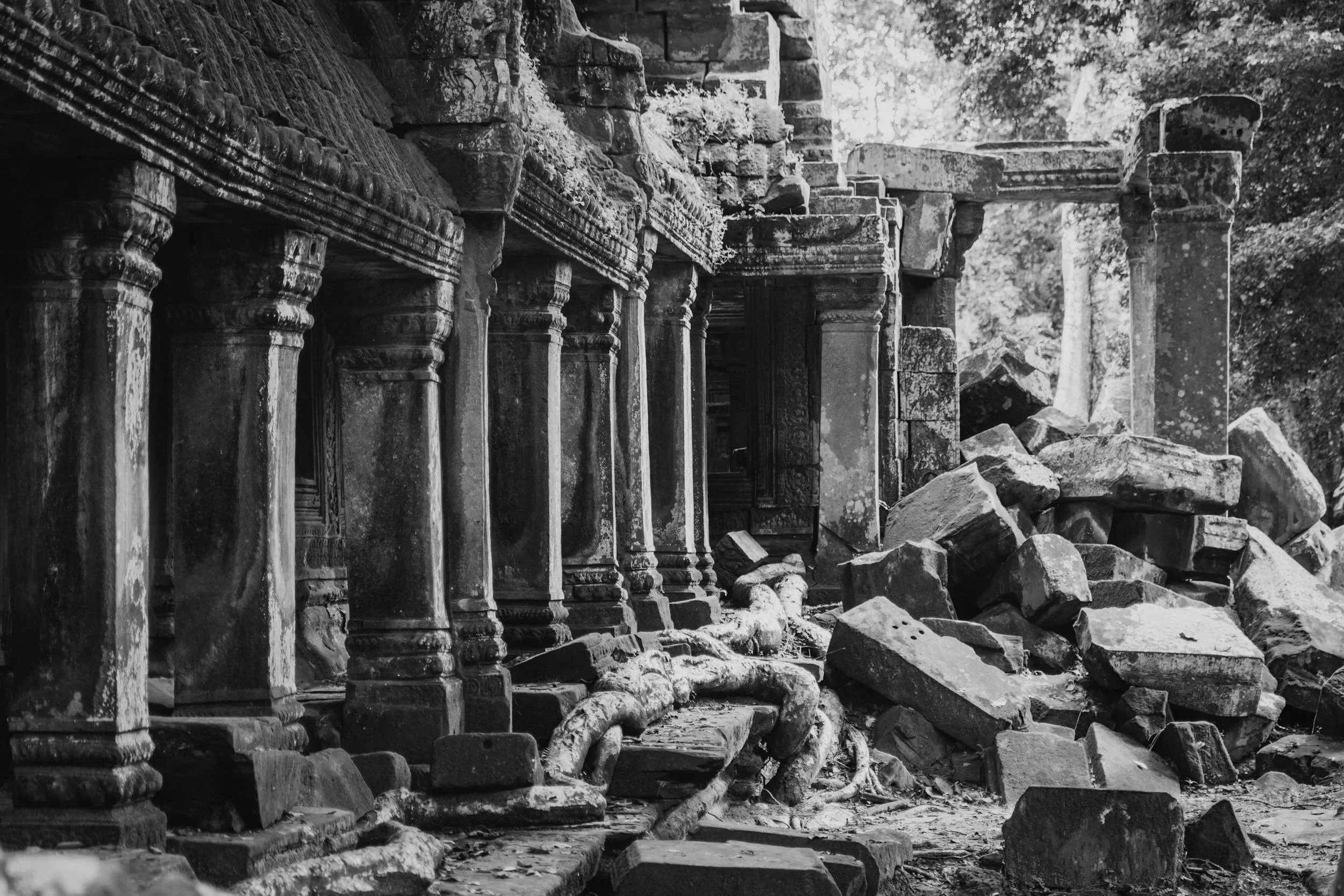 Ancient stone temple ruins with intricately carved columns, some fallen and scattered on the ground, surrounded by dense trees.