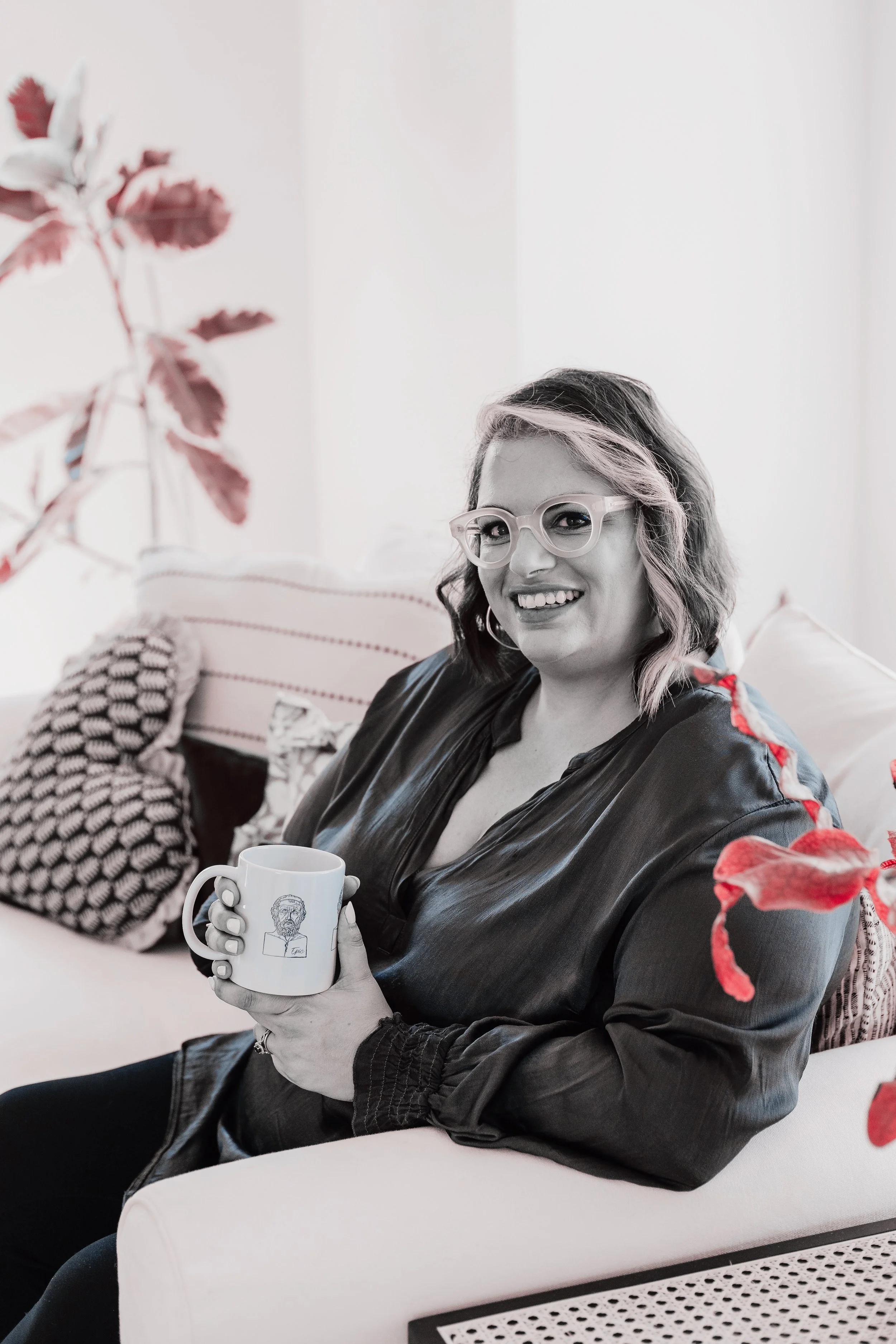 A woman with wavy hair and glasses, wearing a black satin blazer, smiling and holding a white coffee mug with a sketch of a person's face on it. She is seated on a white sofa with decorative pillows and is in a bright room with white walls and colorful plants.
