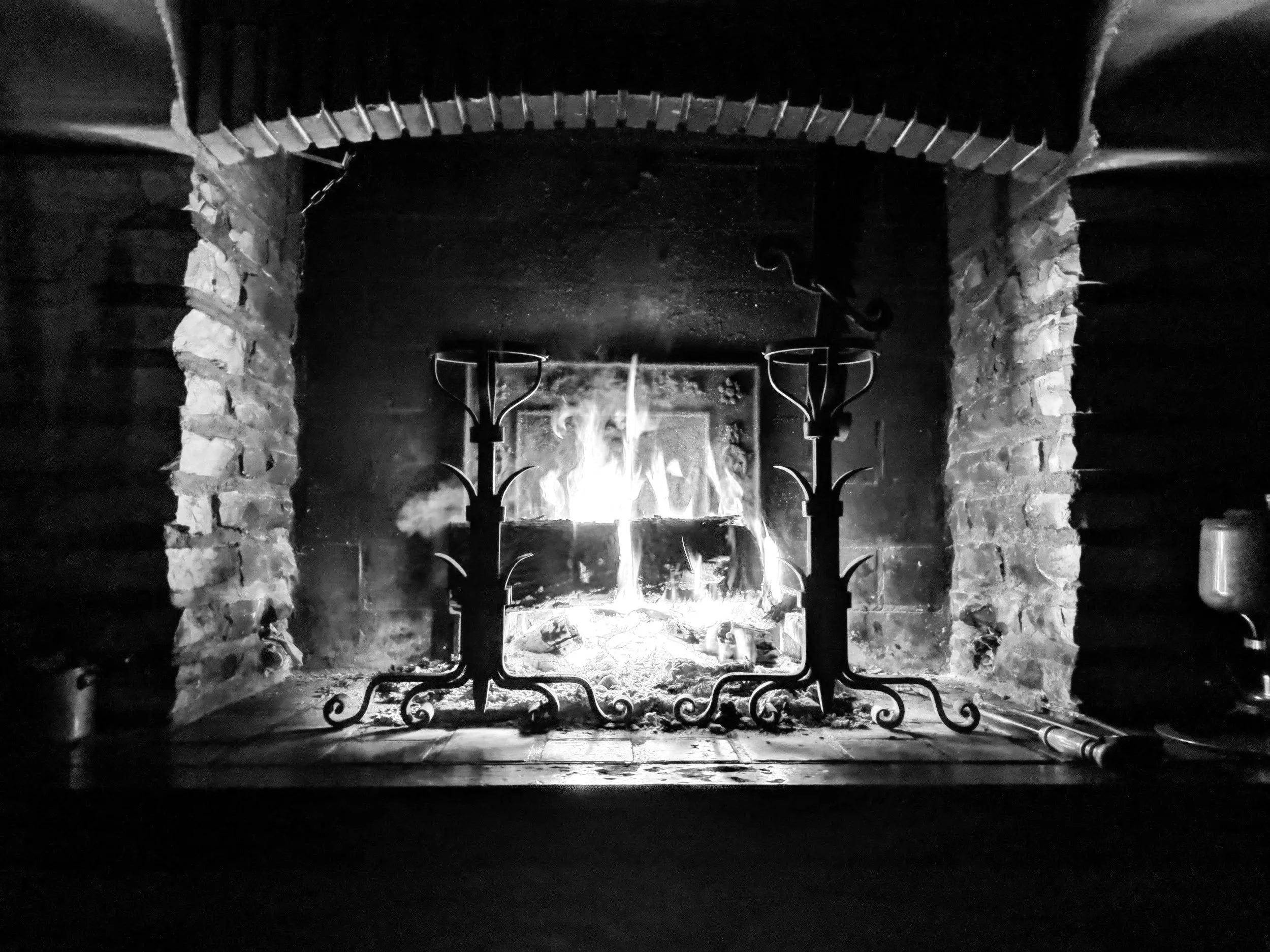 A black and white photo of a stone fireplace with a fire burning inside. There are two candelabra-style holders in front of the fireplace, each with multiple candle holders, but no candles in them.