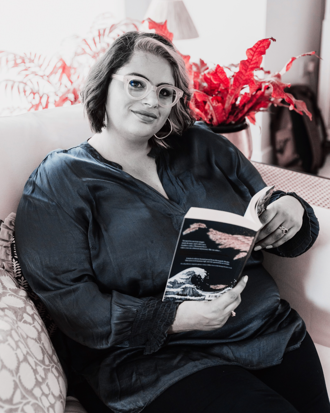 A woman with short hair, glasses, and hoop earrings sitting on a couch, holding a book, with a poinsettia plant in the background.
