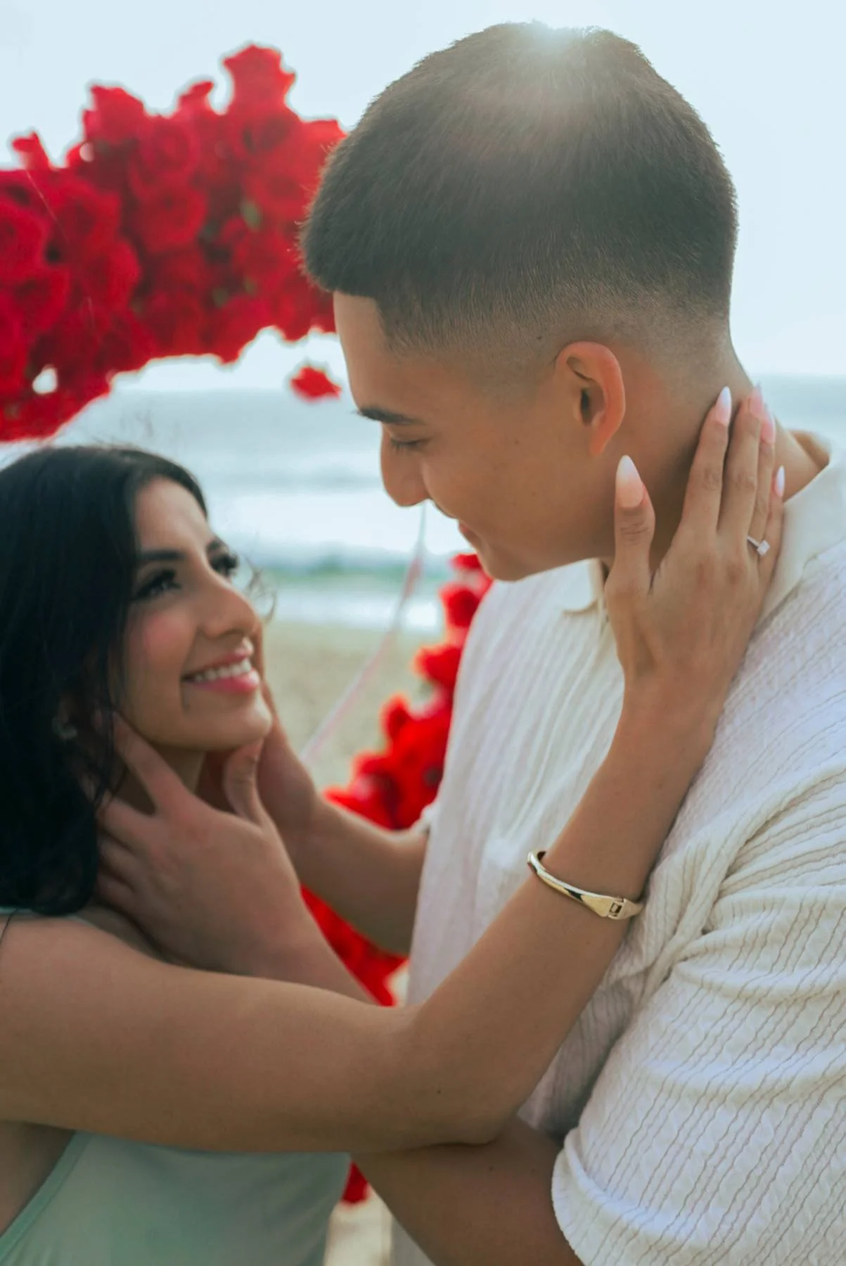 A couple on the beach during daytime, affectionately looking at each other with the woman gently holding the man's face and the man holding the woman's face with both hands, standing in front of a red floral backdrop.