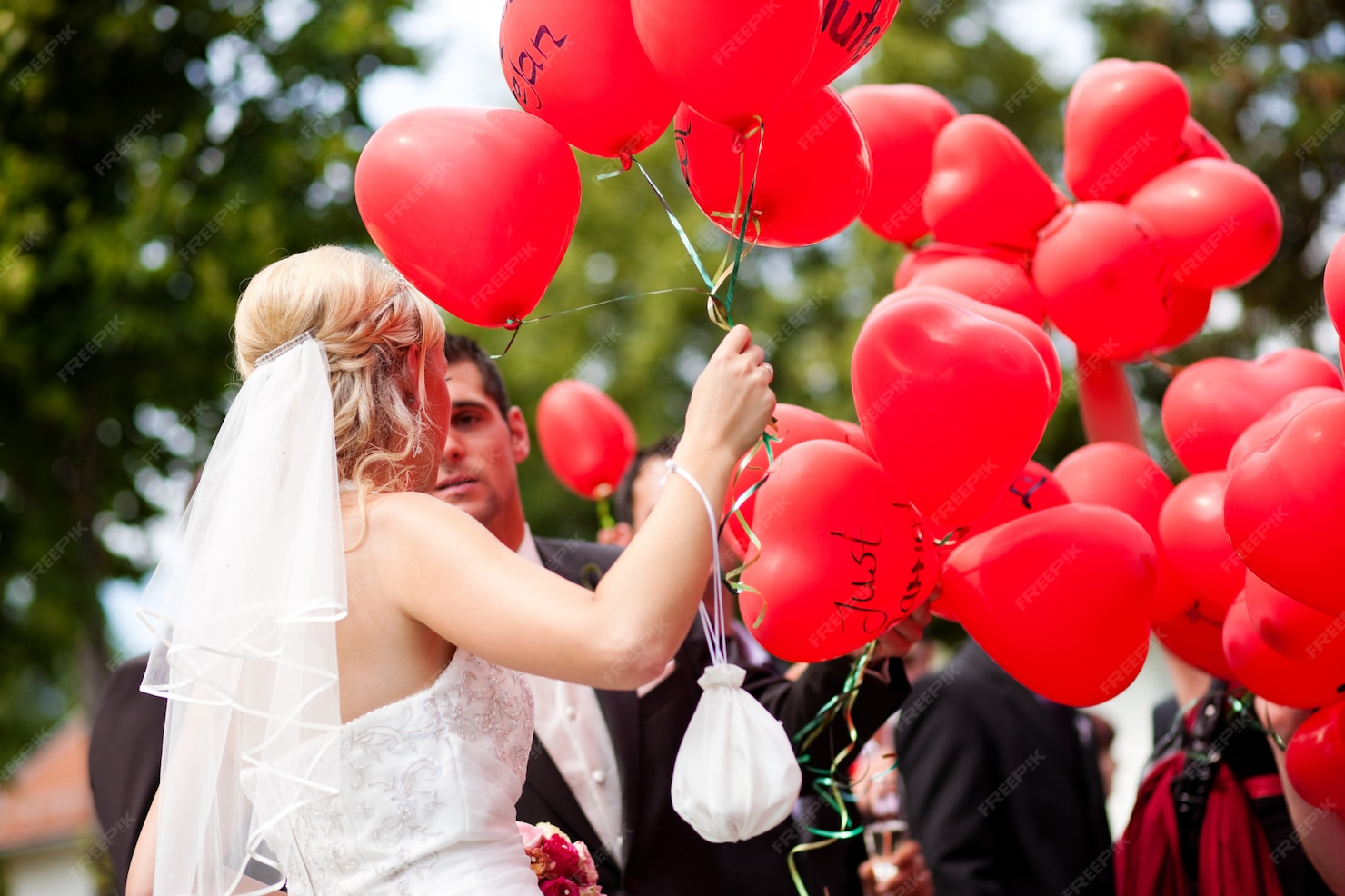 Bride and groom holding red heart-shaped balloons during a wedding ceremony outdoors.