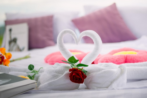 A bed decorated with towel swans forming a heart, with a red rose, pink heart-shaped towels, and purple pillows in the background.