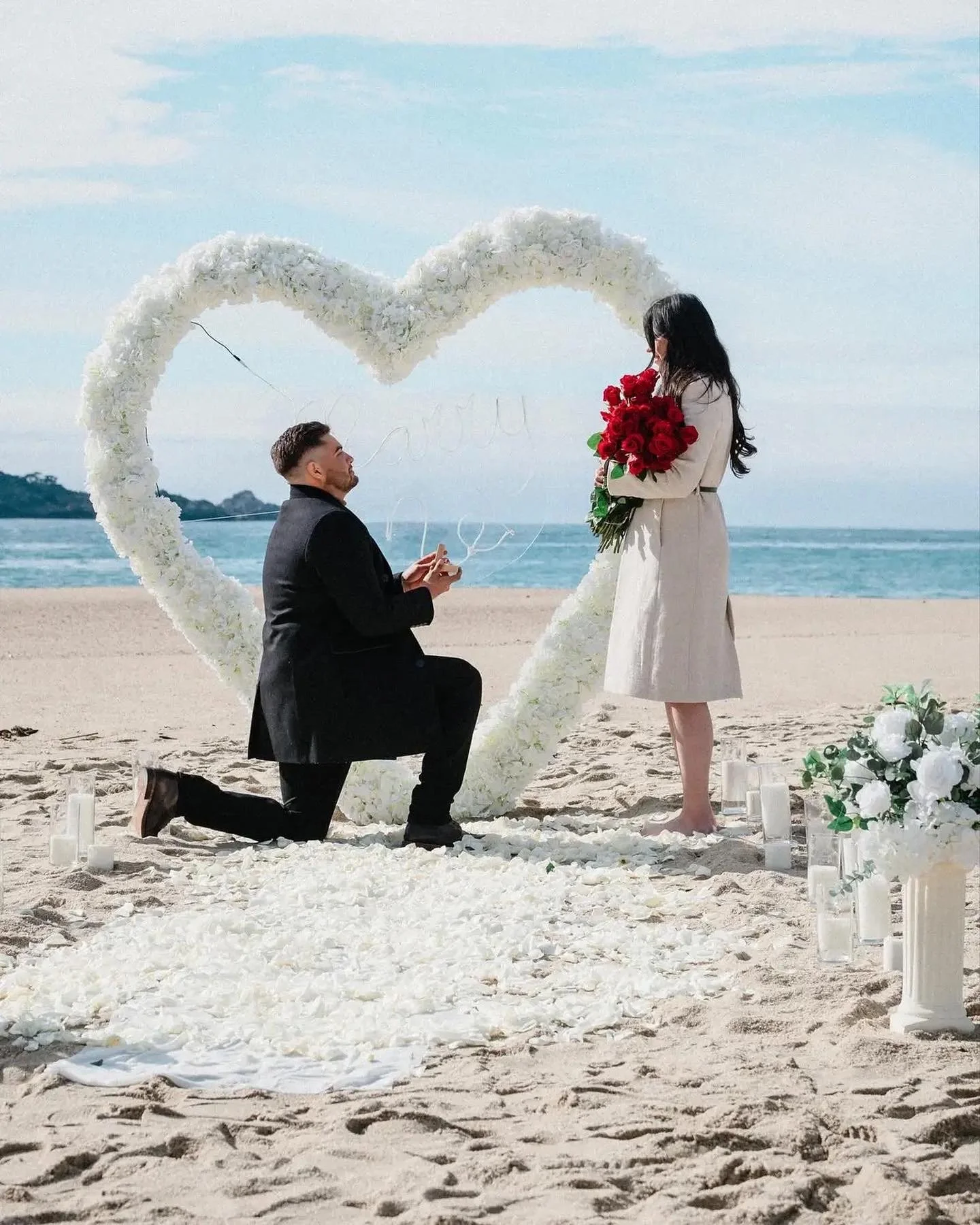 Man proposing marriage to woman on a beach with a heart-shaped floral arch and white flowers along the aisle.
