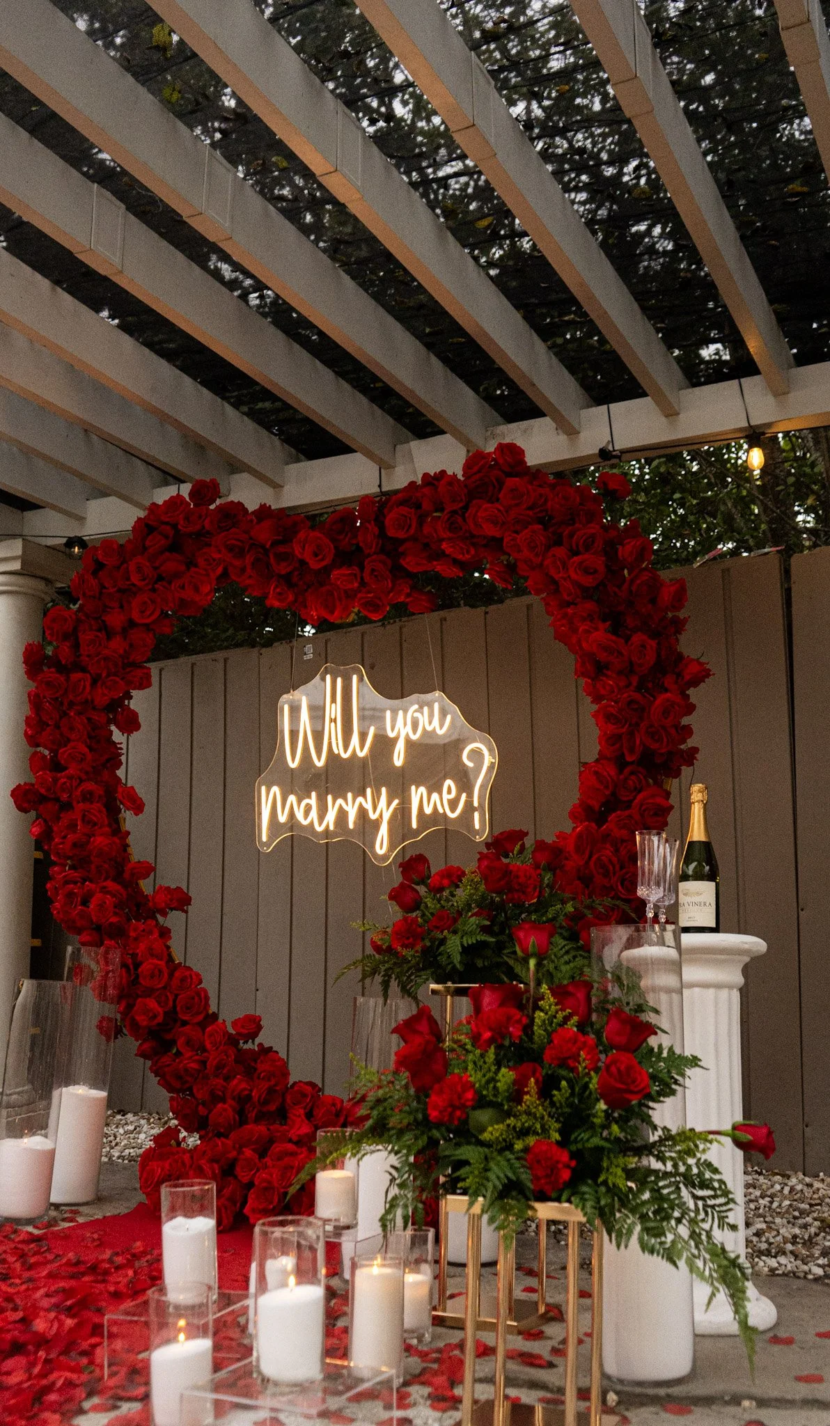 A romantic wedding proposal setup with a large heart-shaped floral arrangement made of red roses, a sign glowing with the words 'Will you marry me?', candles, champagne, and floral decorations.