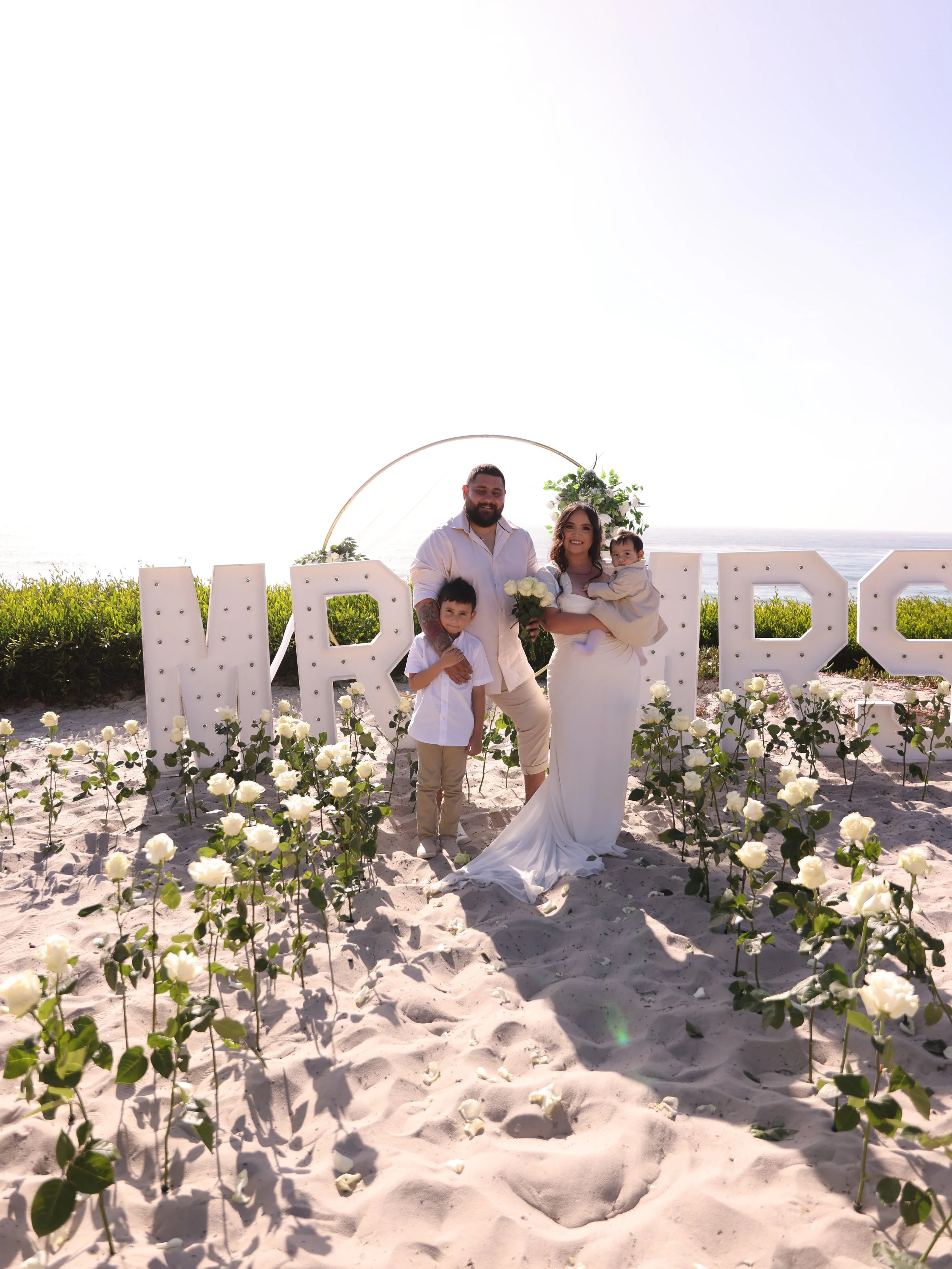 A family celebrating a wedding on a beach with large illuminated letters spelling 'MR' in the background, surrounded by white roses.