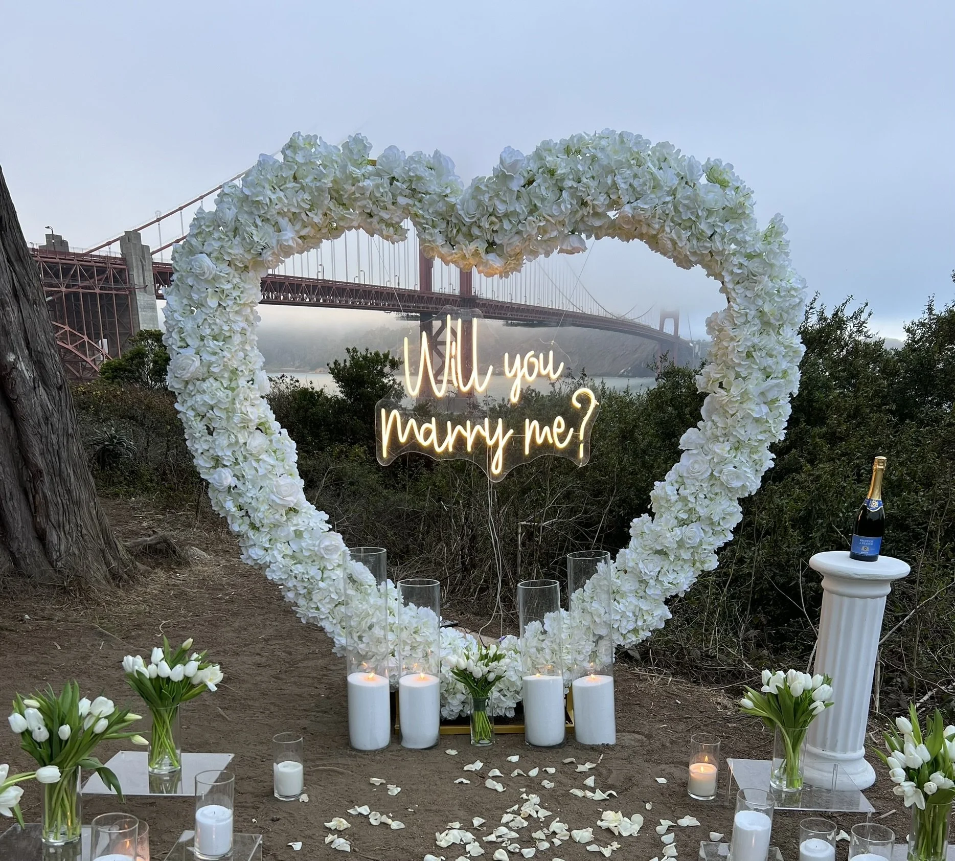 A large white heart-shaped floral arch with a neon sign reading 'Will you marry me?' in front of the Golden Gate Bridge. The setup includes white candles, white flowers in glass vases, and a bottle of champagne on a white pedestal, with rose petals s