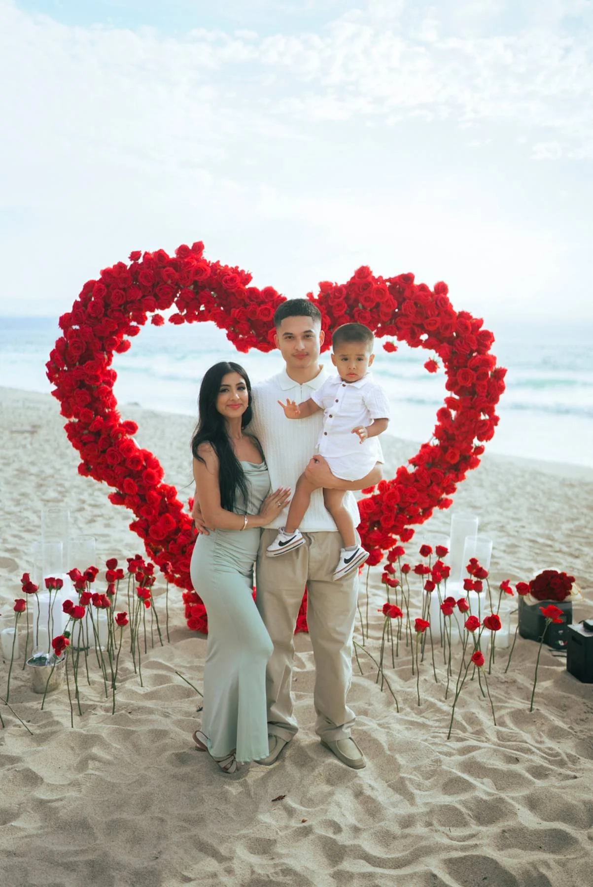 A family of three stands on a sandy beach in front of a large heart-shaped arrangement of red roses. The woman is wearing a light-colored dress, and the man is dressed in light-colored pants and a short-sleeved shirt. The young boy, held by the man, 