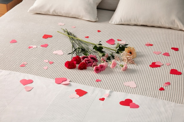 Bouquet of mixed flowers on bed with scattered paper hearts.