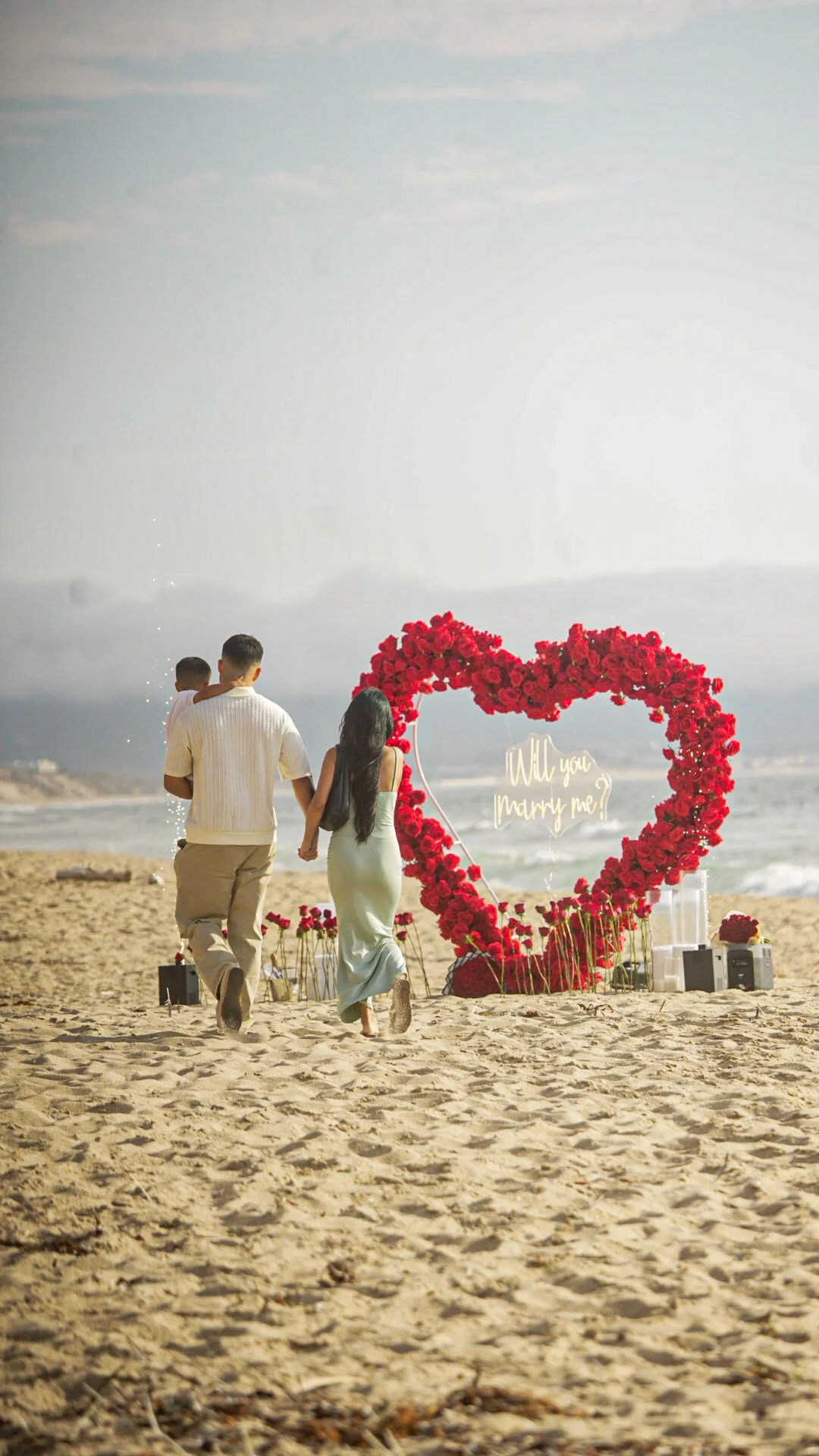 A couple holding hands walking on a sandy beach towards a rose heart-shaped arch with a sign that says 'Will you marry me?' in the background.