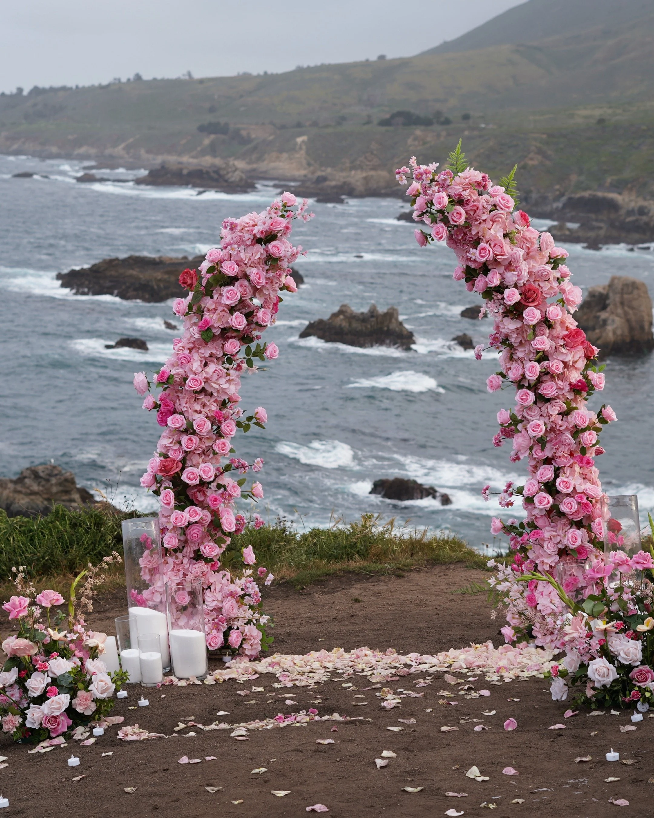 A floral arch made of pink roses and greenery on a dirt path, overlooking the ocean with rocks and a hilly coastline in the background, decorated with candles and flower petals.