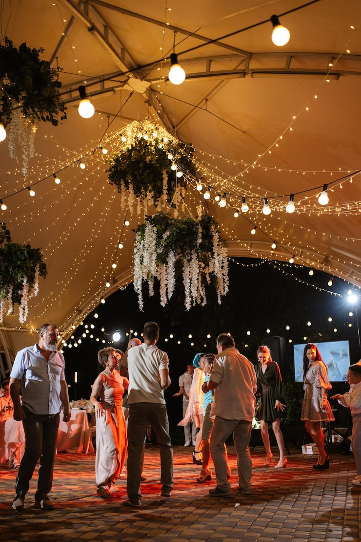People dancing under a decorated tent with hanging string lights and floral arrangements at night.