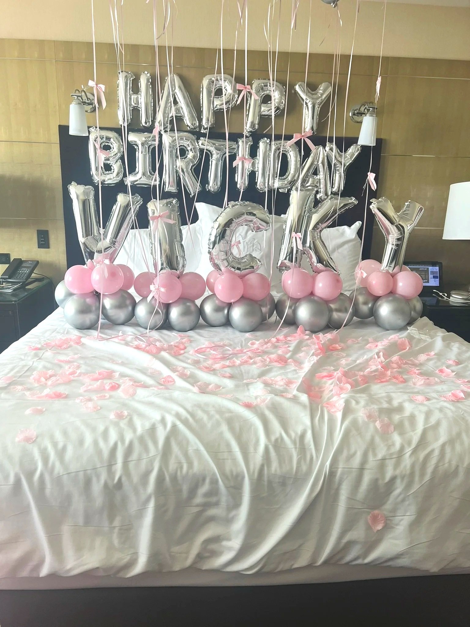 Decorated bed with pink and silver balloons spelling out 'Happy Birthday VICKY', pink ribbons, and rose petals.