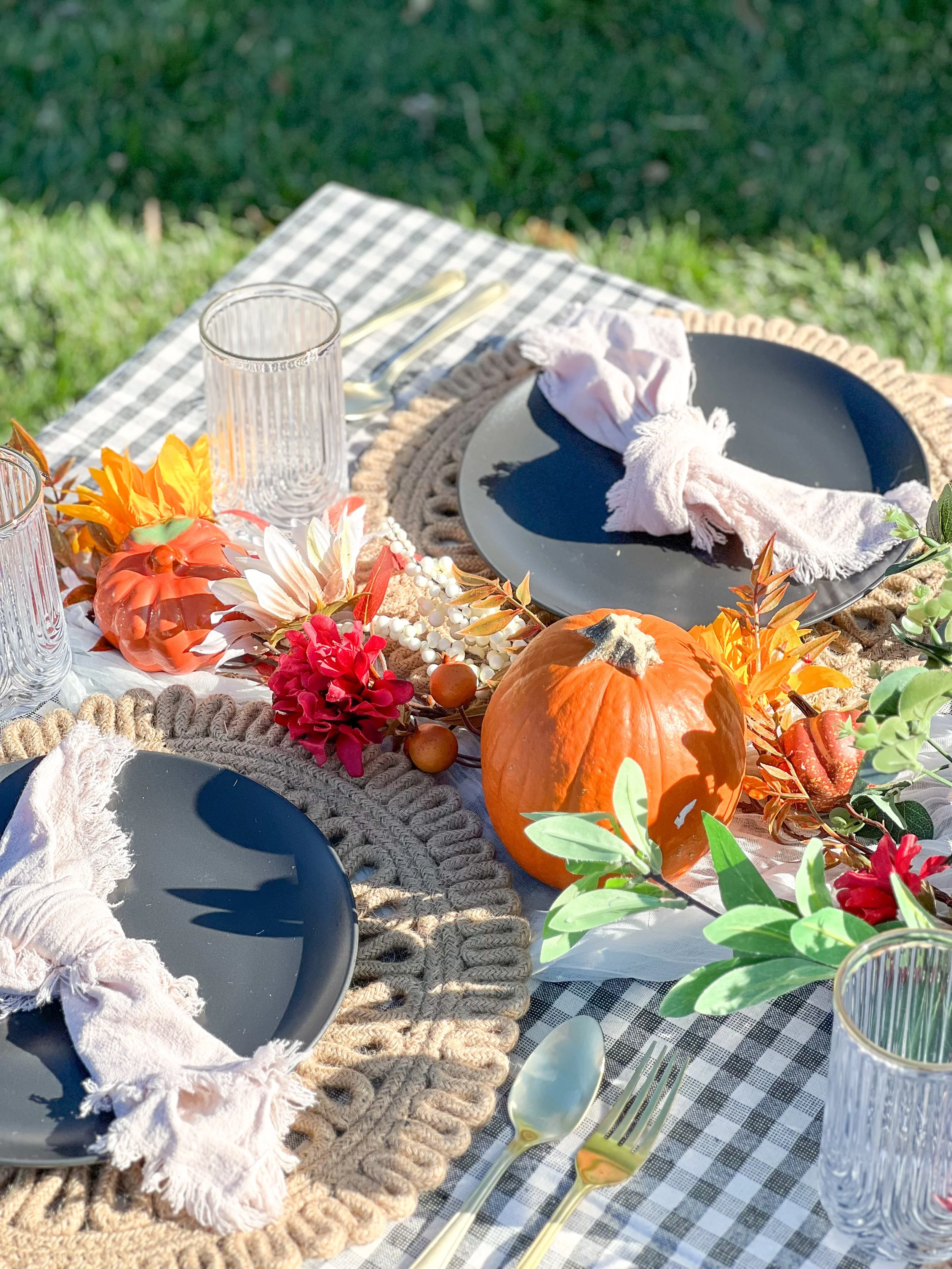 Fall-themed outdoor dining table with black plates, pink napkins, glassware, and a pumpkin centerpiece, decorated with autumn leaves and flowers.