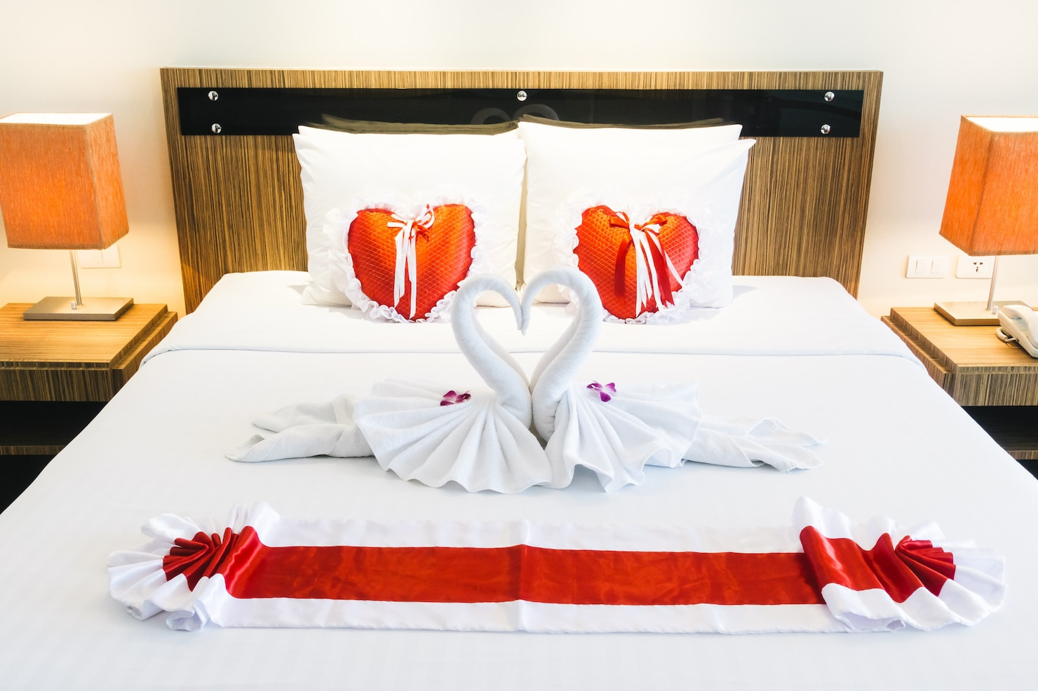 A hotel bed decorated with two towel swans forming a heart, red and white pillow hearts, purple flower petals, and a red and white fabric runner, with side tables, lamps, and a telephone.