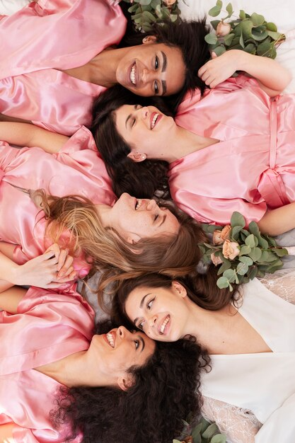 Five women lying down together on a soft surface, wearing satin robes in pink and white, surrounded by eucalyptus and peach roses, all smiling and looking at each other.